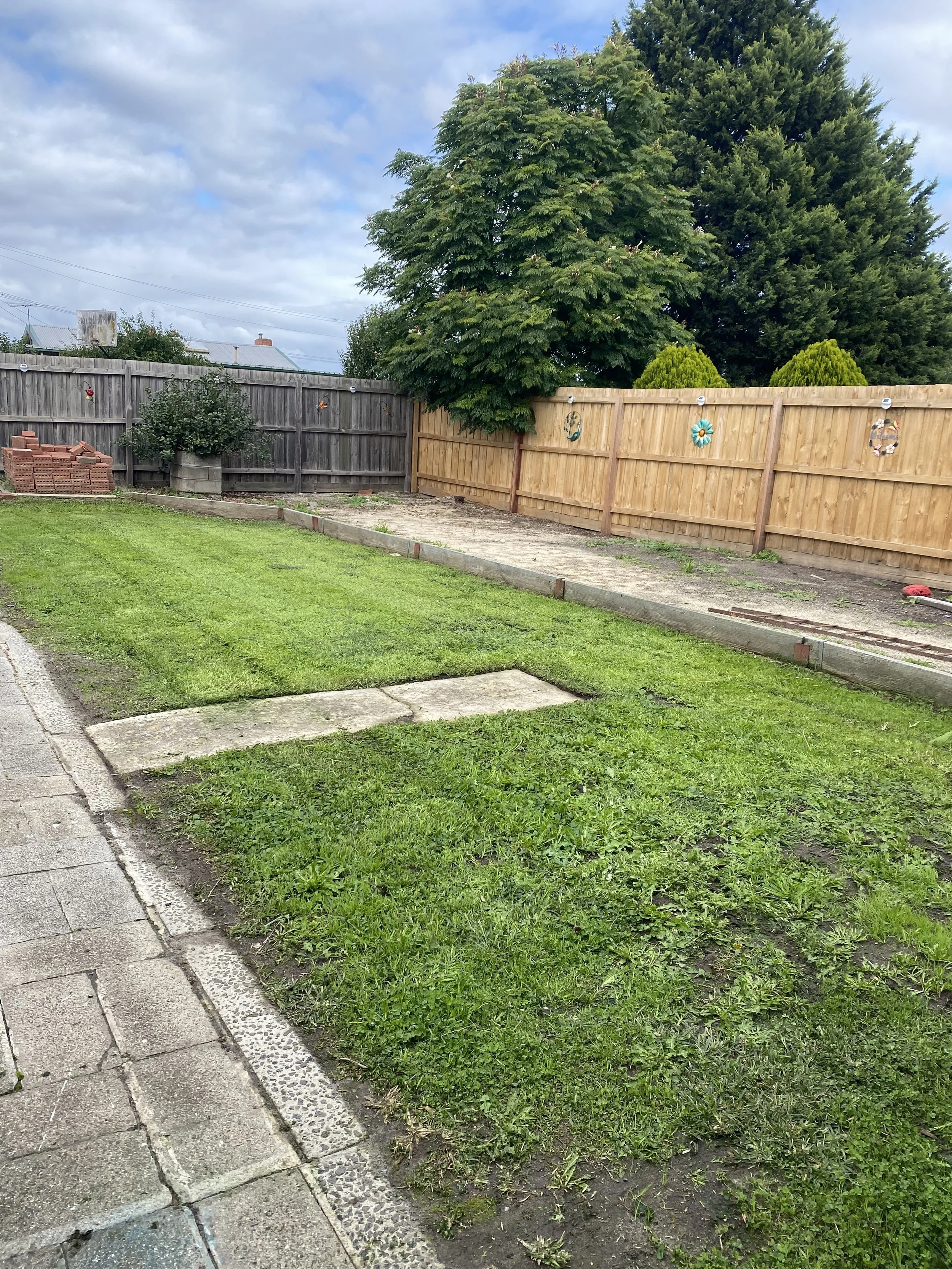 The image shows a small backyard with a freshly built wooden fence on the right side, decorated with ceramic garden ornaments. There is a lush green lawn in the foreground, with a sidewalk and curb at the left edge. A corner of the yard has some soil and a few bricks stacked nearby. In the background, there are large trees, one with broad green leaves and the other a darker evergreen. The sky is partly cloudy with patches of blue.