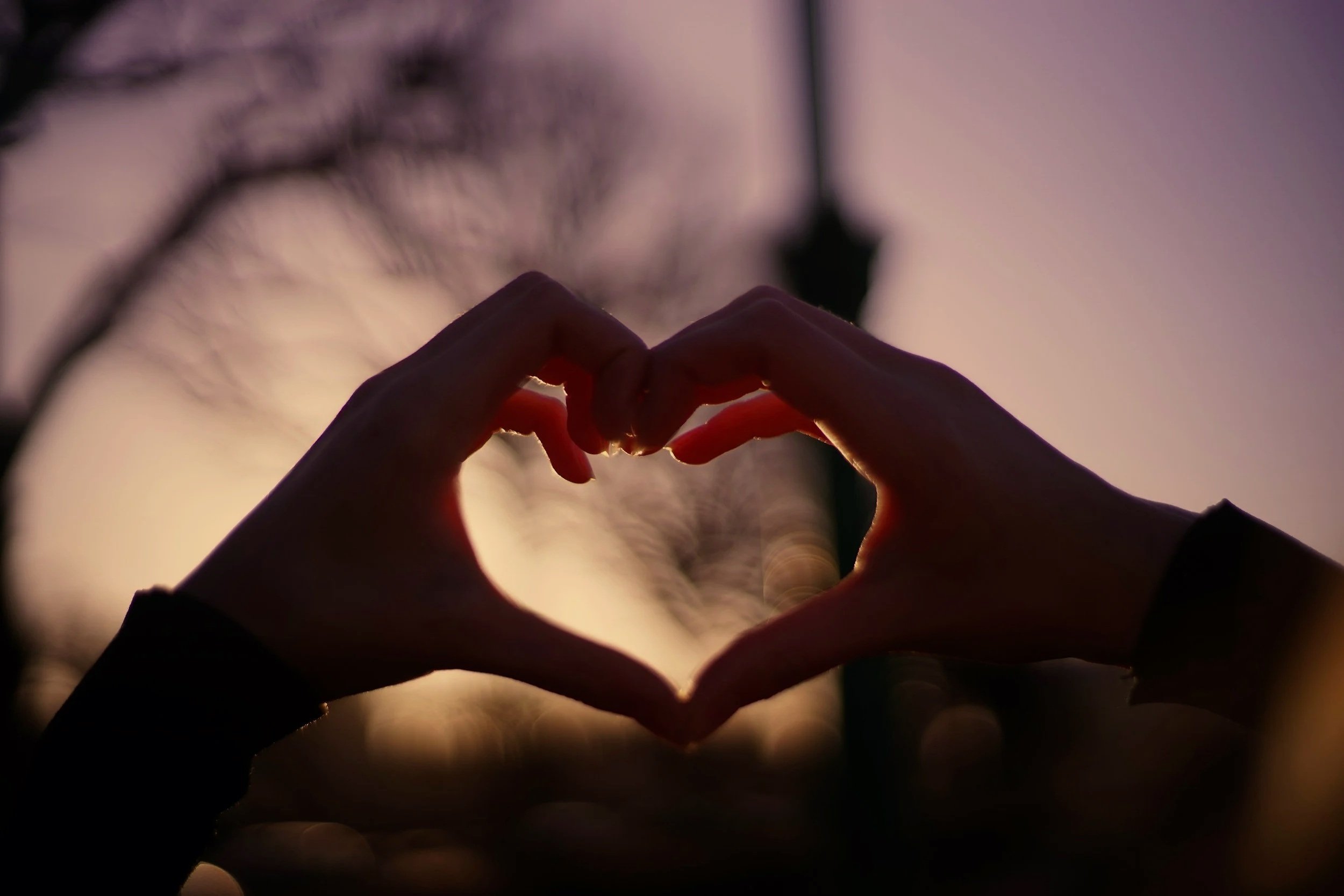 Two hands forming a heart shape with fingers at sunset, with the sky in the background.