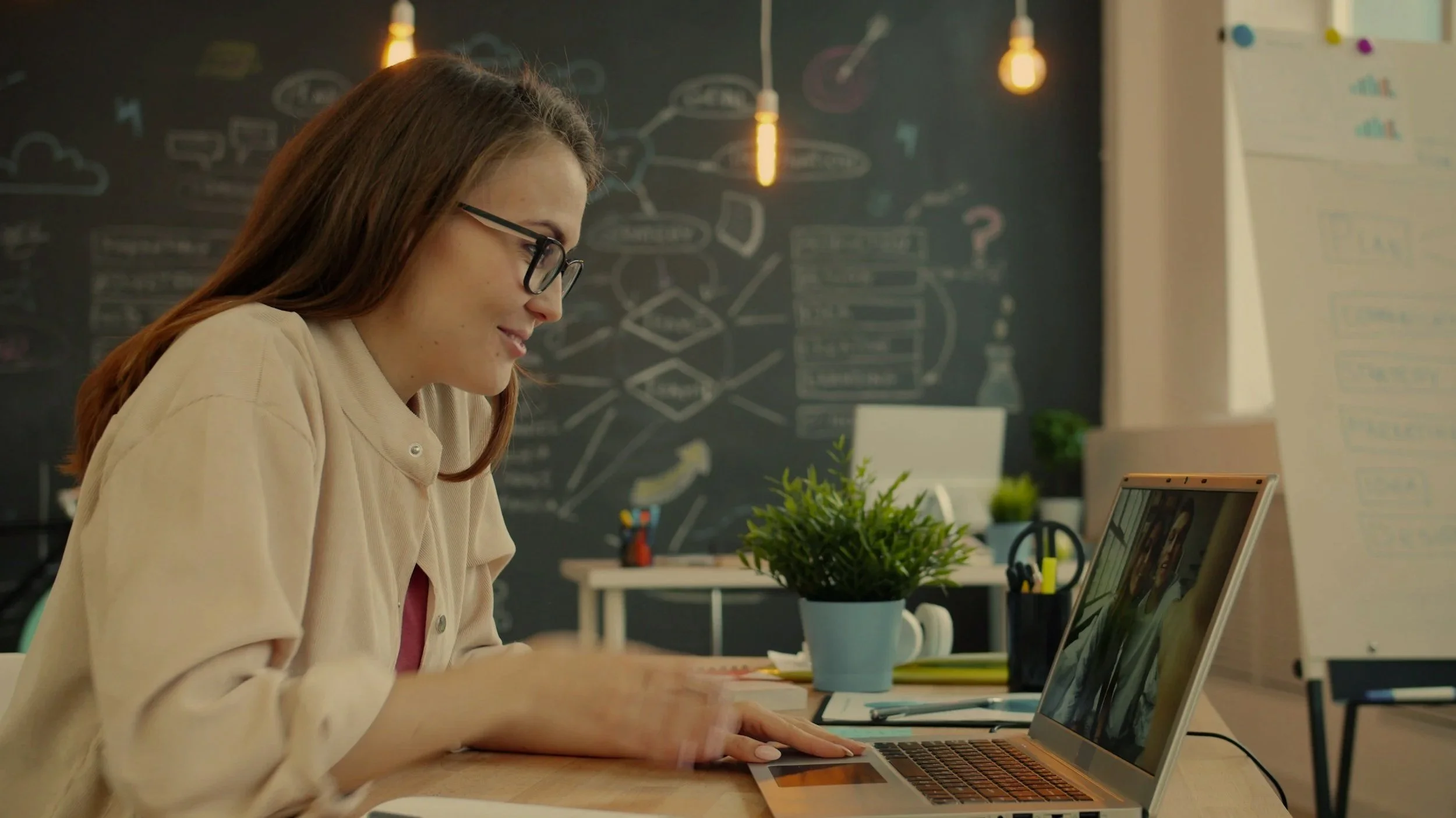 A woman with glasses is sitting at a desk, working on a laptop in a modern office. The background has a black chalkboard wall with various drawings and notes, hanging light bulbs, and a whiteboard with colorful charts.