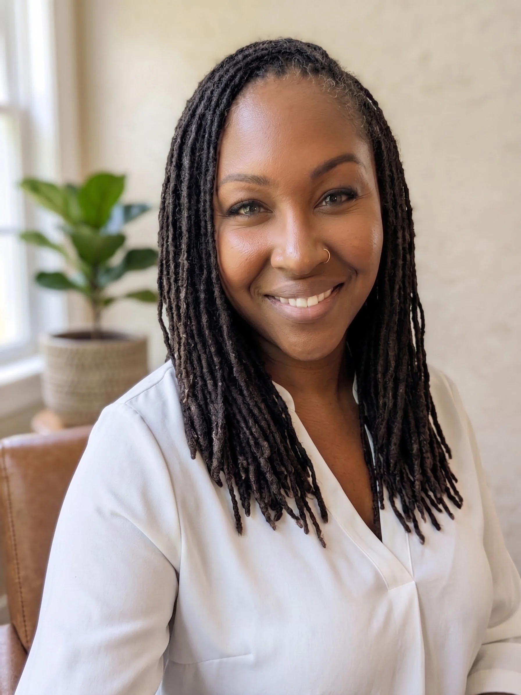 Close-up of a woman with long, dark dreadlocks, smiling and wearing a white top, sitting in a room with a beige wall and a green potted plant in the background.