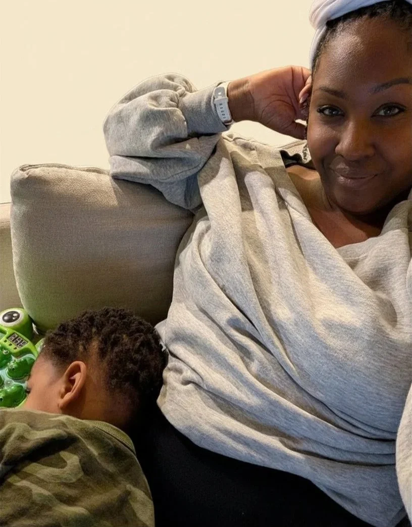 A woman relaxing on a beige sofa with a young boy sleeping next to her, wearing a camouflage shirt, and a green toy nearby.