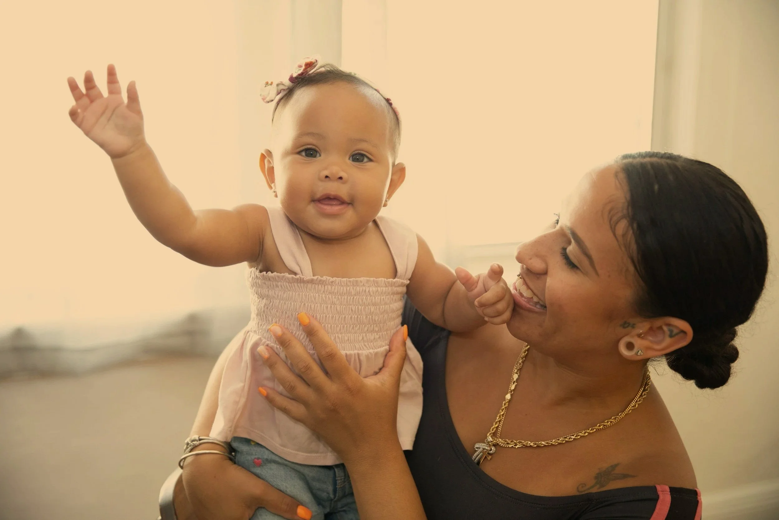 A woman holding a smiling baby girl, who is waving, in a bright room with cream-colored curtains.