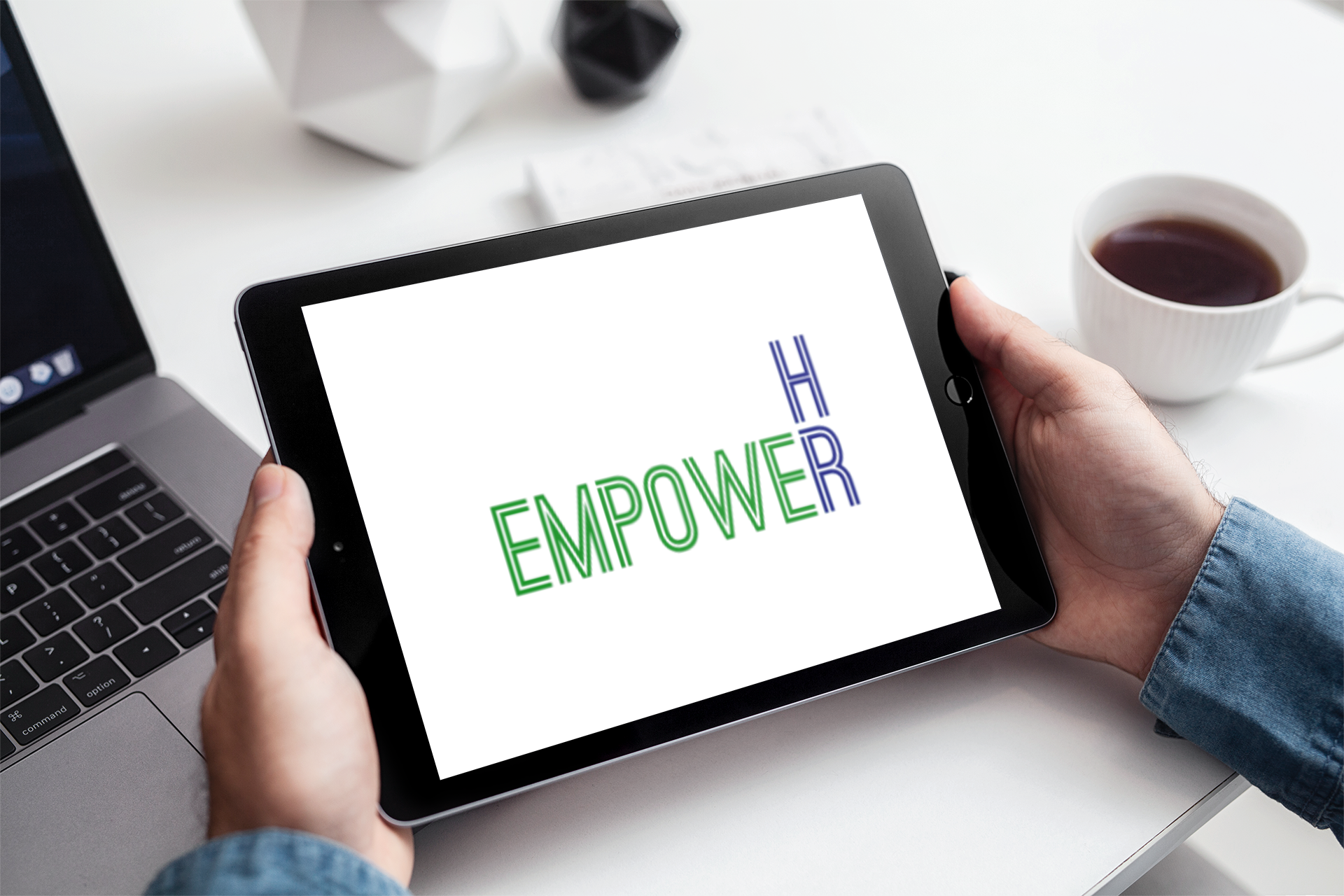 Person holding a tablet displaying the words 'BE EMPOWER' in colorful text, on a white desk with a laptop, a coffee cup, and geometric decorative objects.