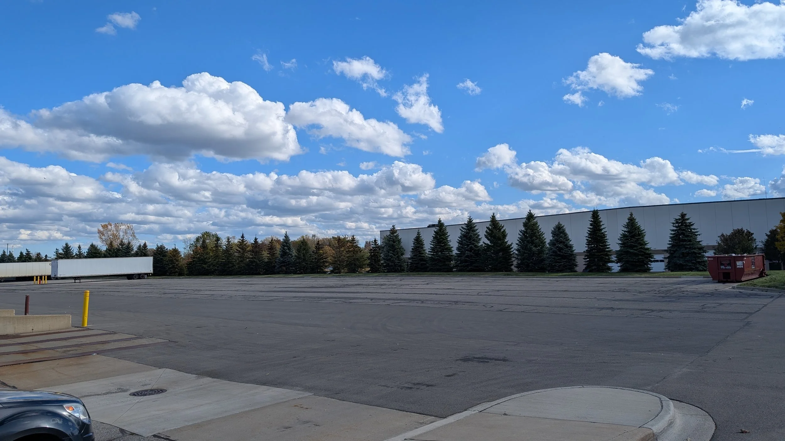 Empty parking lot with a few parked cars, rows of evergreen trees along the background, large warehouse building, blue sky with scattered clouds.