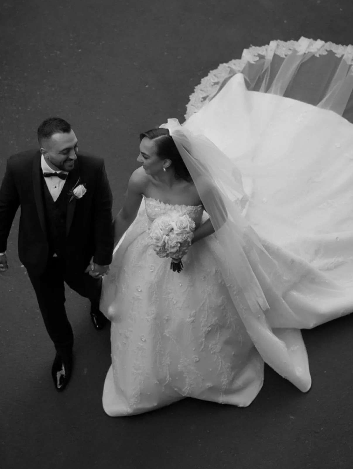 A black-and-white photo of a bride and groom walking hand in hand, smiling, with the bride holding a bouquet of roses, dressed in a wedding gown with a large train, and the groom wearing a tuxedo with a bow tie.
