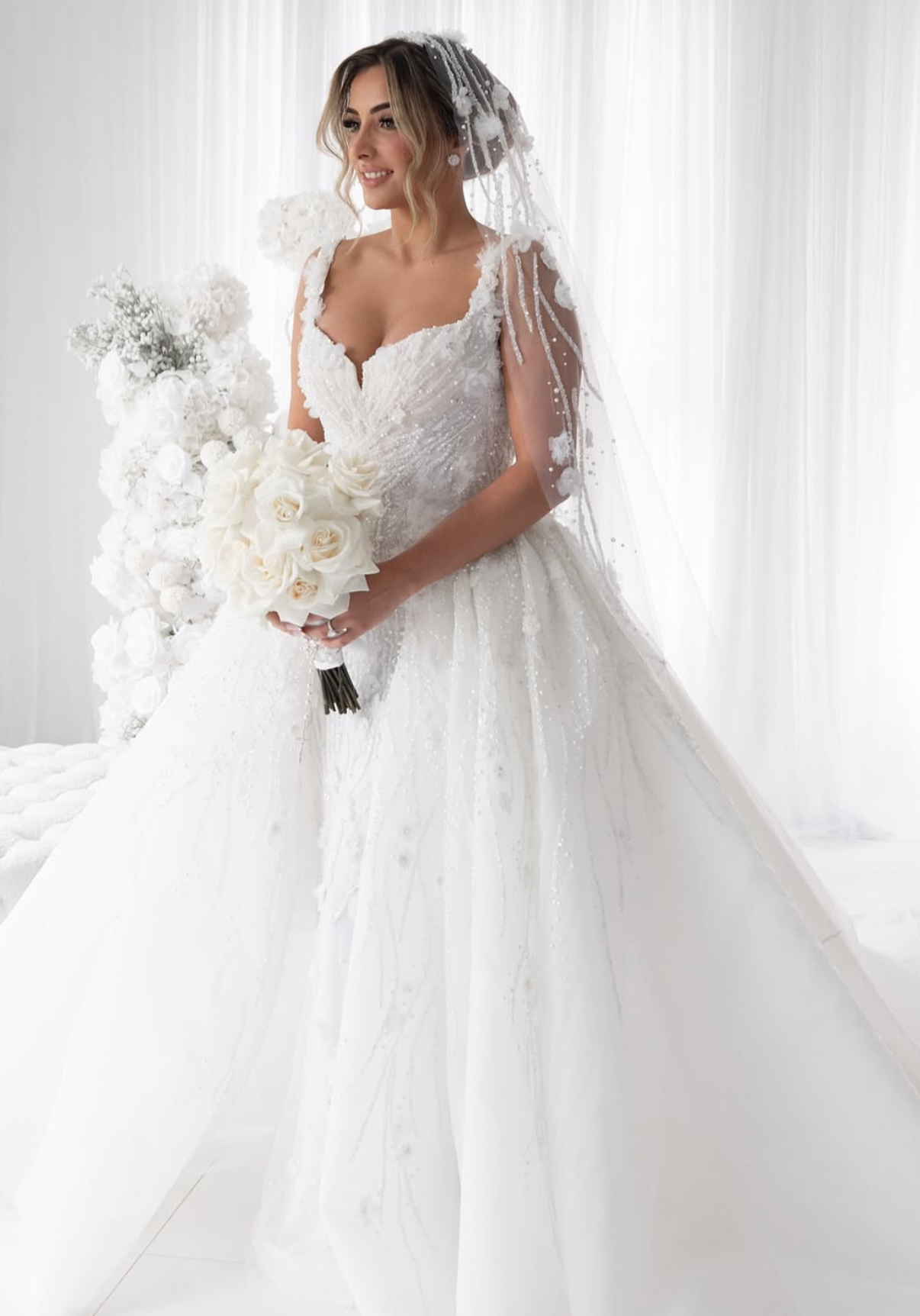 A bride in a white wedding gown holding a bouquet of white roses in a room with white curtains.