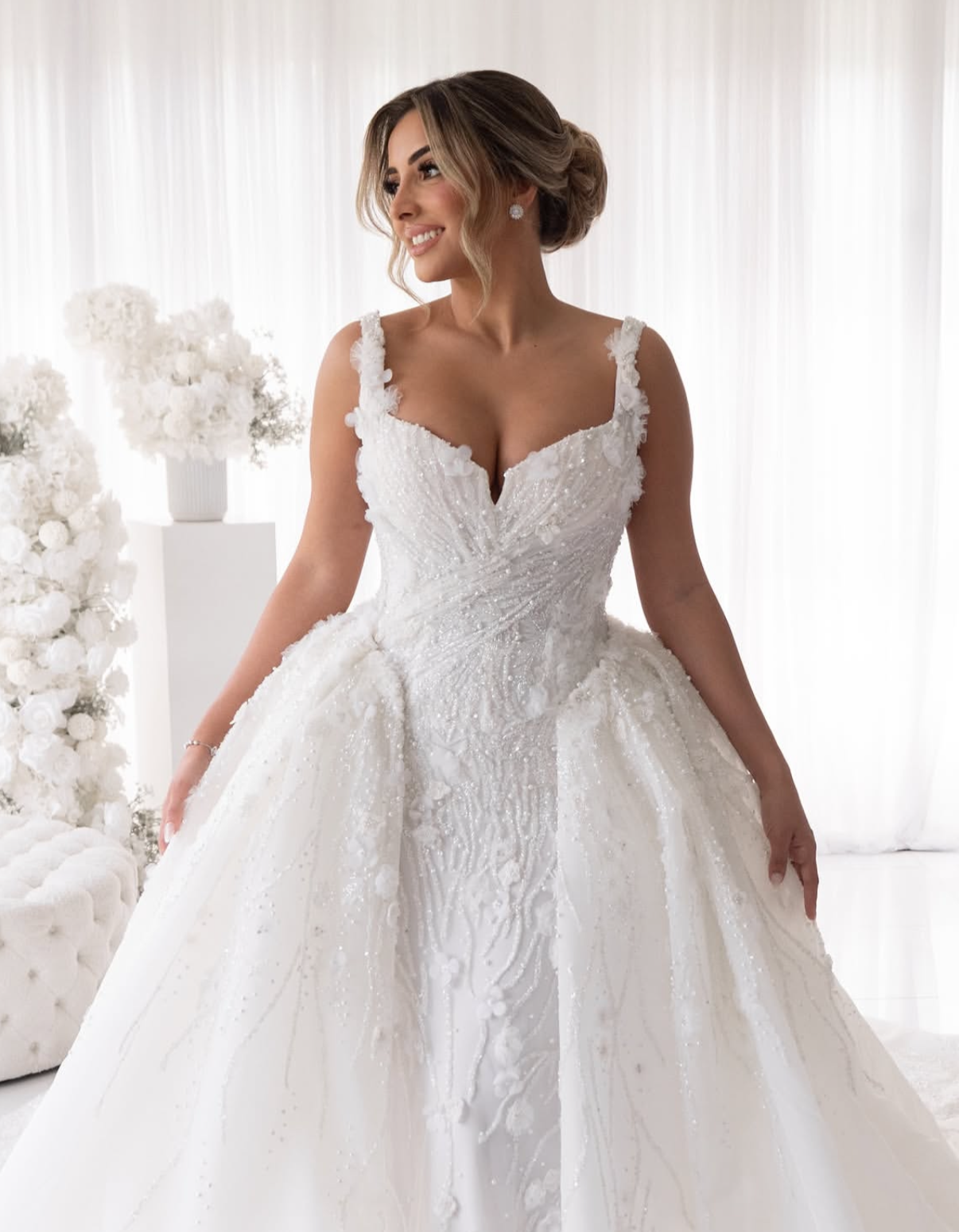 A woman in a wedding dress standing in a bright room with white curtains and floral arrangements.