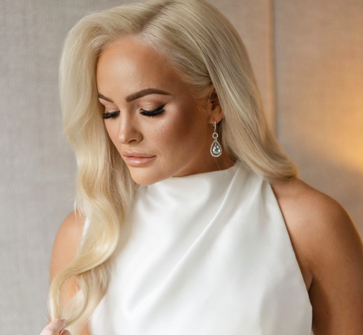 A woman with long blonde hair, makeup, and earrings wearing a white satin sleeveless top, looking down with closed eyes.