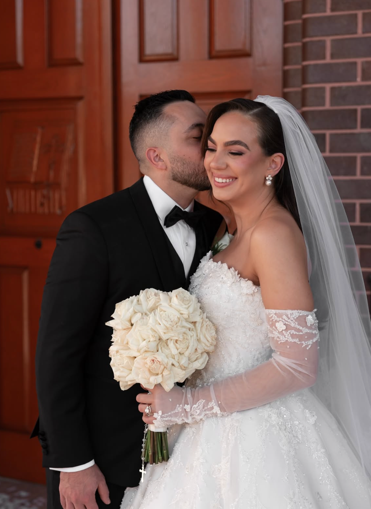 A bride and groom at their wedding, with the groom whispering into the bride's ear. The bride is holding a bouquet of white roses and is smiling. They are standing in front of a wooden door and brick wall.
