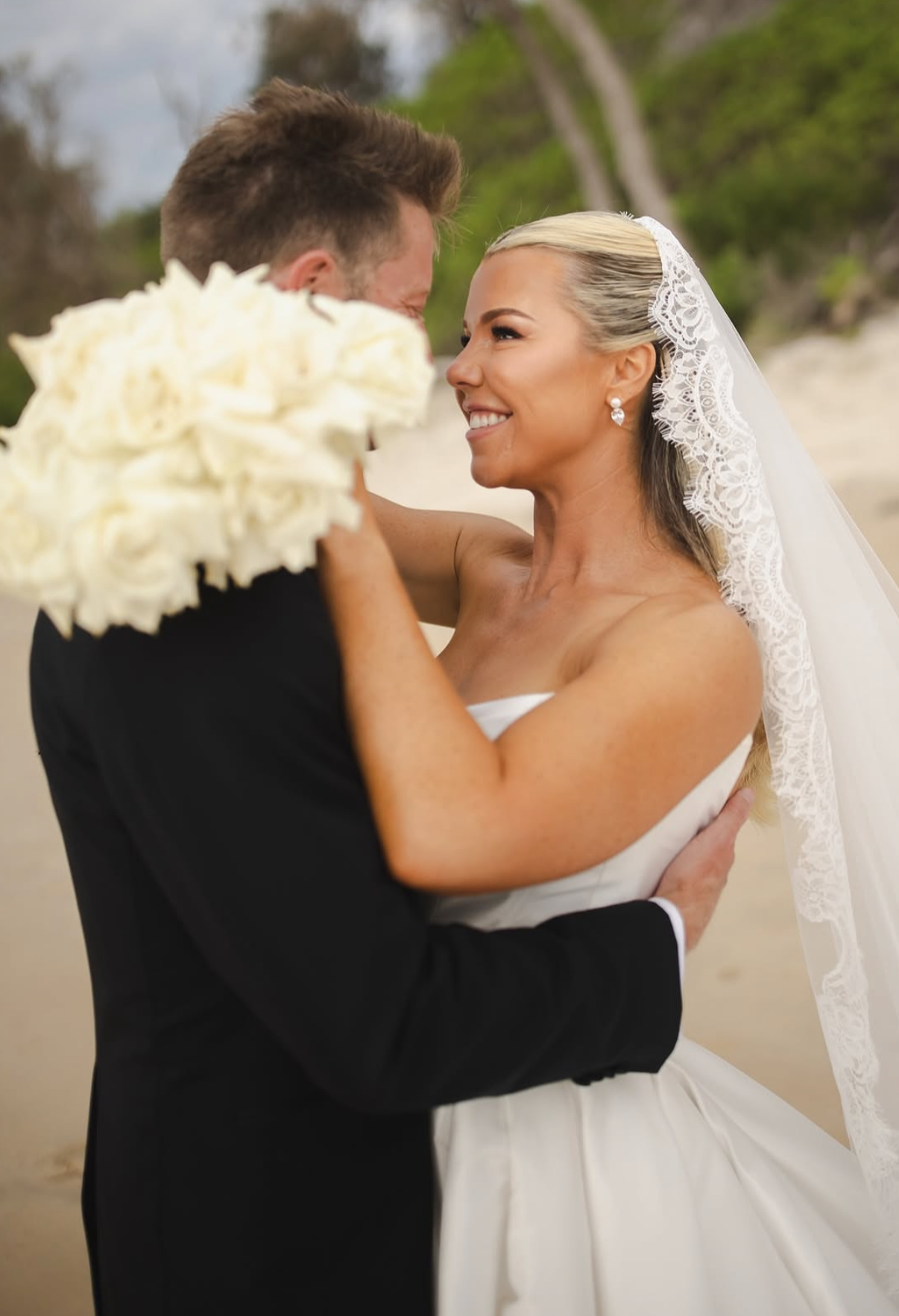 A bride and groom smiling and looking at each other during their outdoor wedding, with the groom holding a bouquet of white flowers.