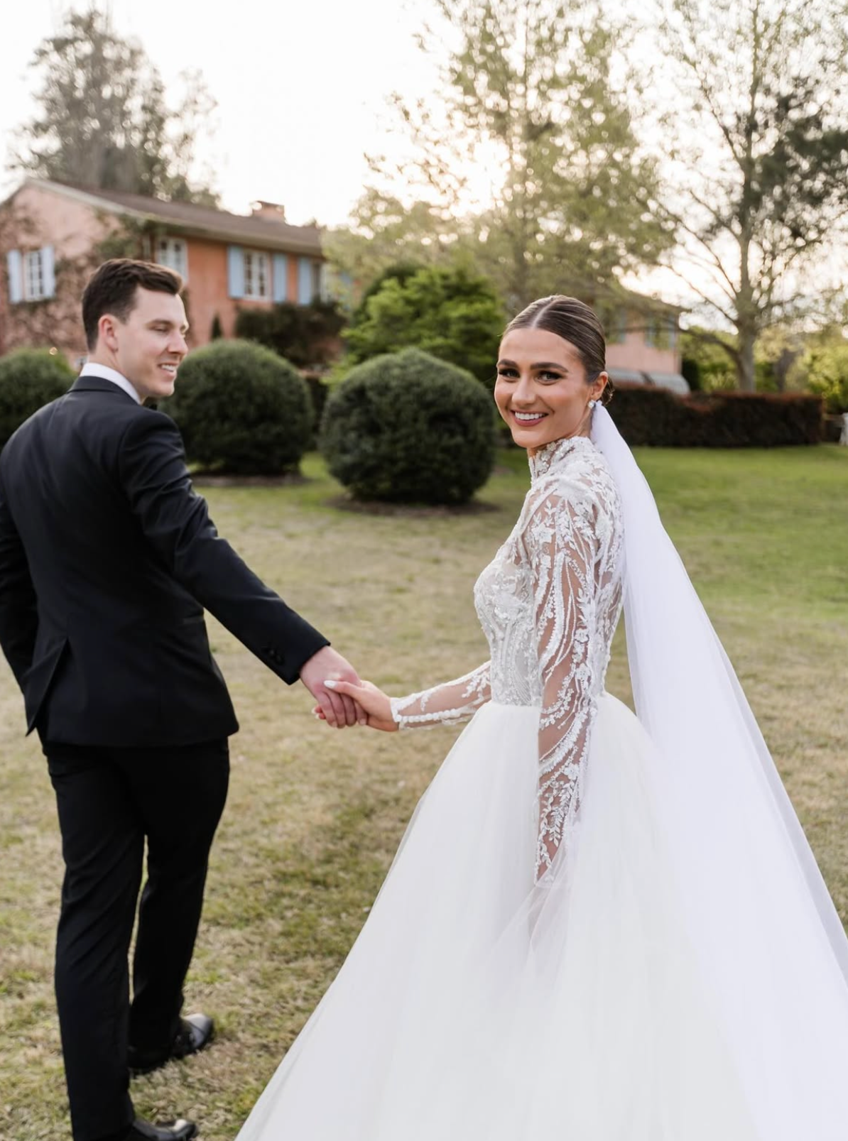 A newlywed couple holding hands outdoors on a grassy lawn, with the bride smiling at the camera and the groom looking at her, surrounded by trees and a house in the background at sunset.