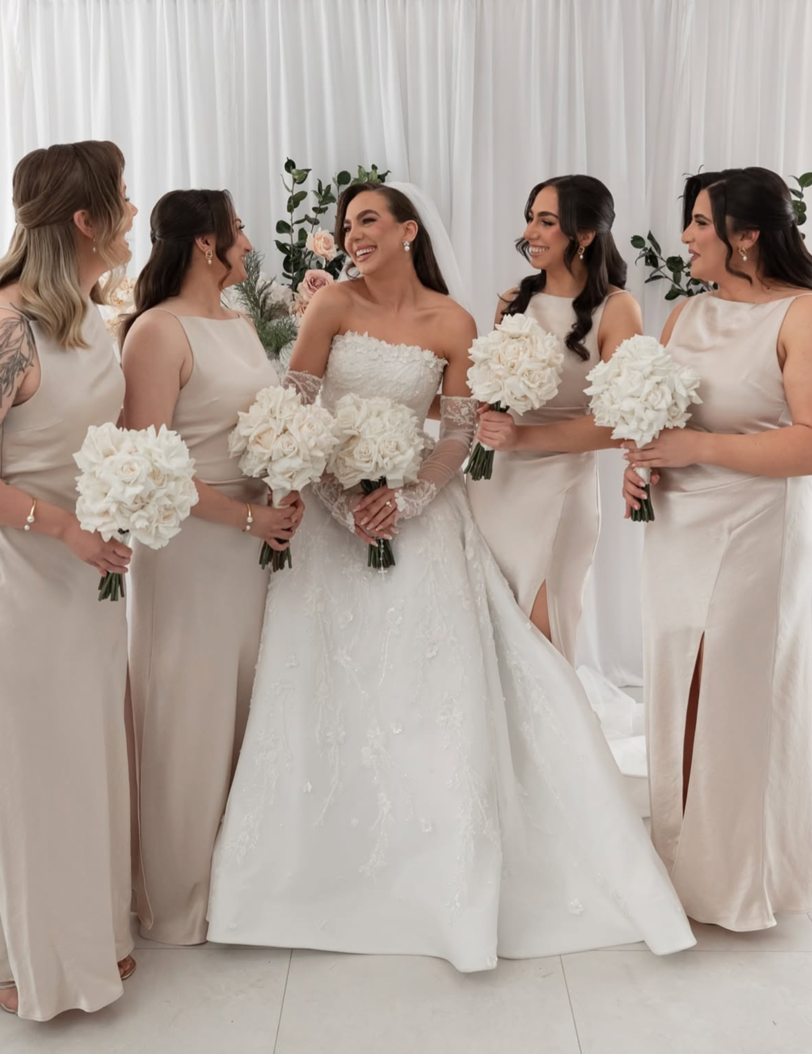 A bride in a white wedding dress surrounded by five bridesmaids in matching cream dresses, all holding white bouquets, standing in front of a white curtain and floral background.