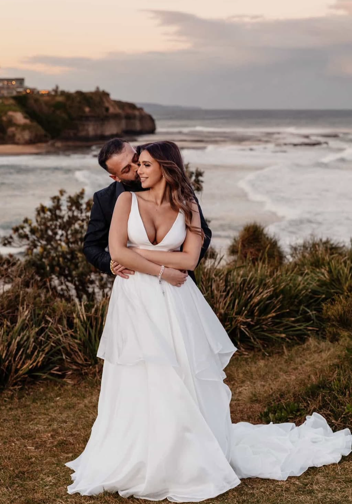 A bride and groom embracing on a beach at sunset, with cliffs and ocean in the background. The bride is in a white wedding gown, and the groom is in a dark suit.