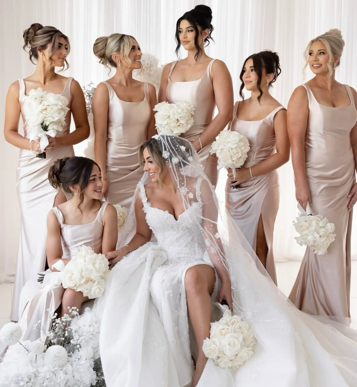 A bride in a white wedding dress with a veil sitting on a chair, surrounded by seven women in beige bridesmaid dresses, all holding white bouquets, in front of a light background.