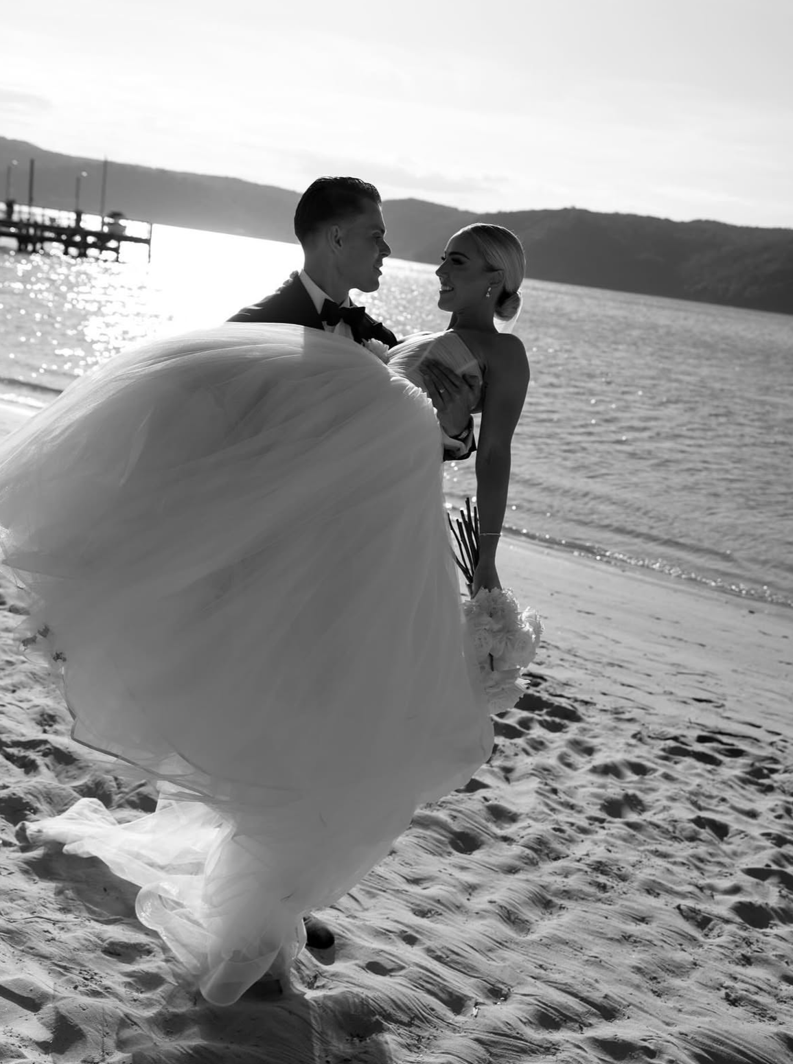 A black and white photo of a newlywed couple on a beach, with the groom holding the bride in his arms. The groom is dressed in a tuxedo, and the bride is wearing a strapless wedding gown. The bride is holding a bouquet of flowers, and they are smilin