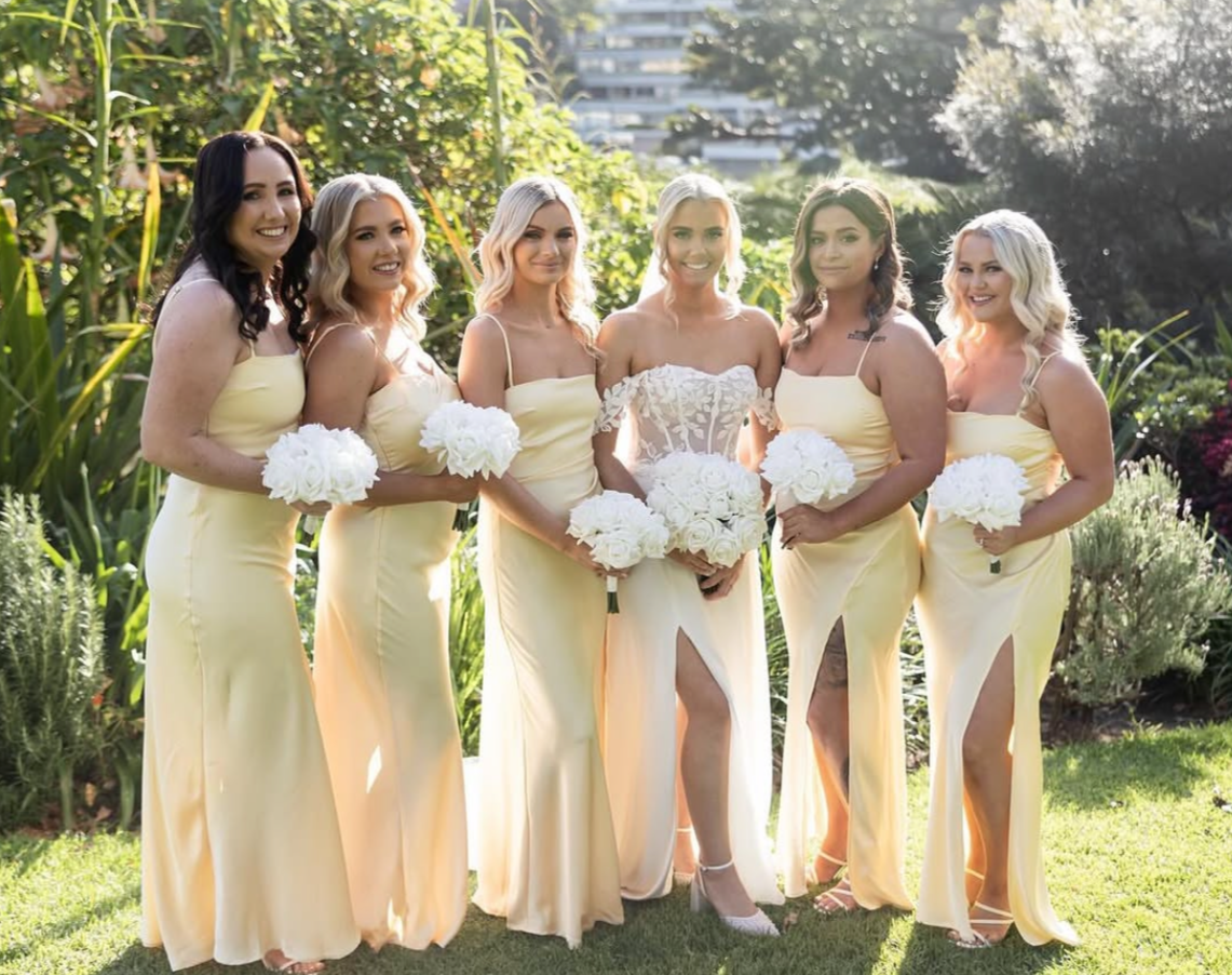 Group of six women in formal dresses, with the bride in the center holding a bouquet of flowers, standing outdoors in a garden with greenery and sunlight.