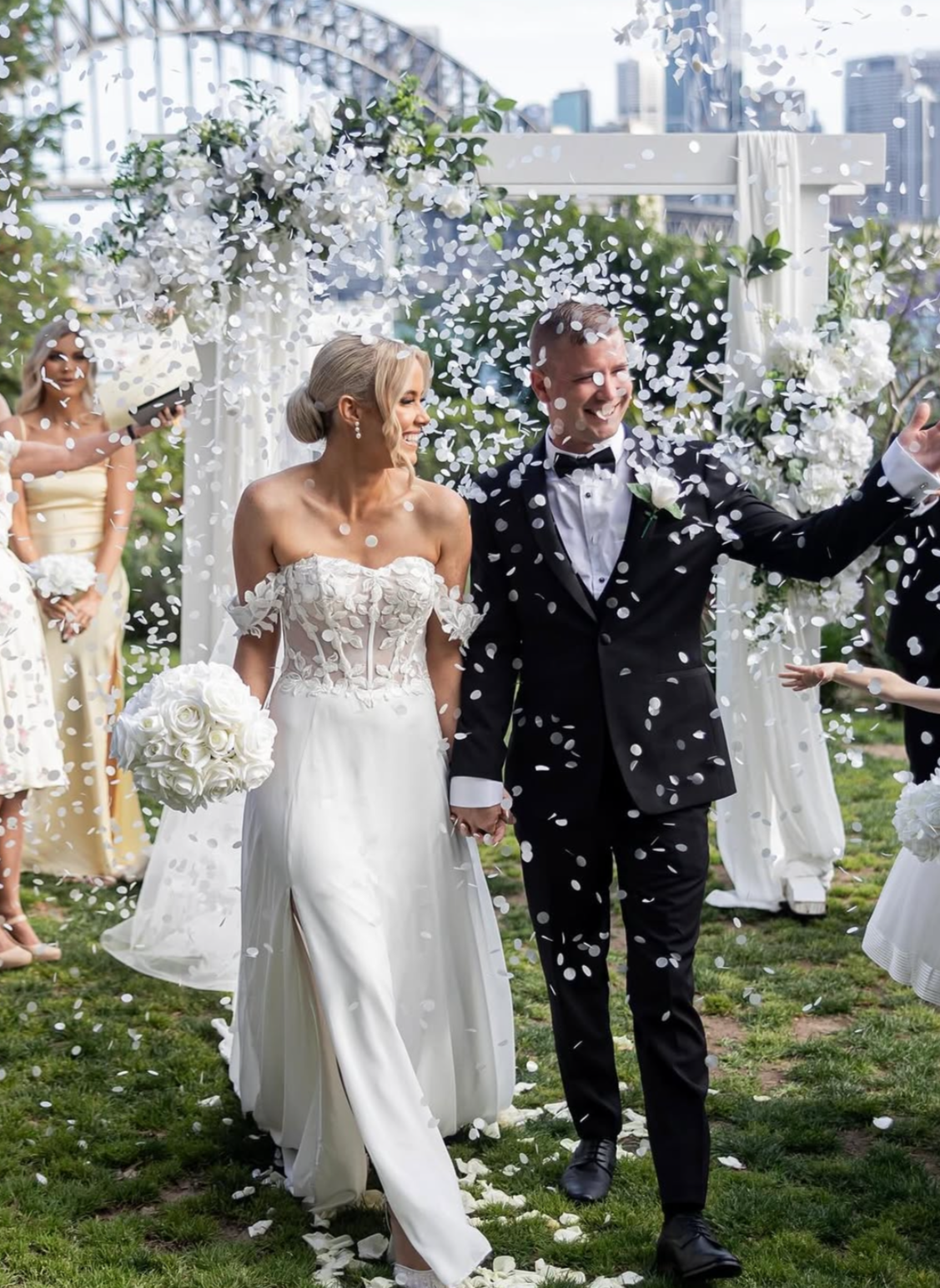 A newlywed couple walks hand-in-hand through falling white confetti at their outdoor wedding ceremony, with a cityscape in the background.