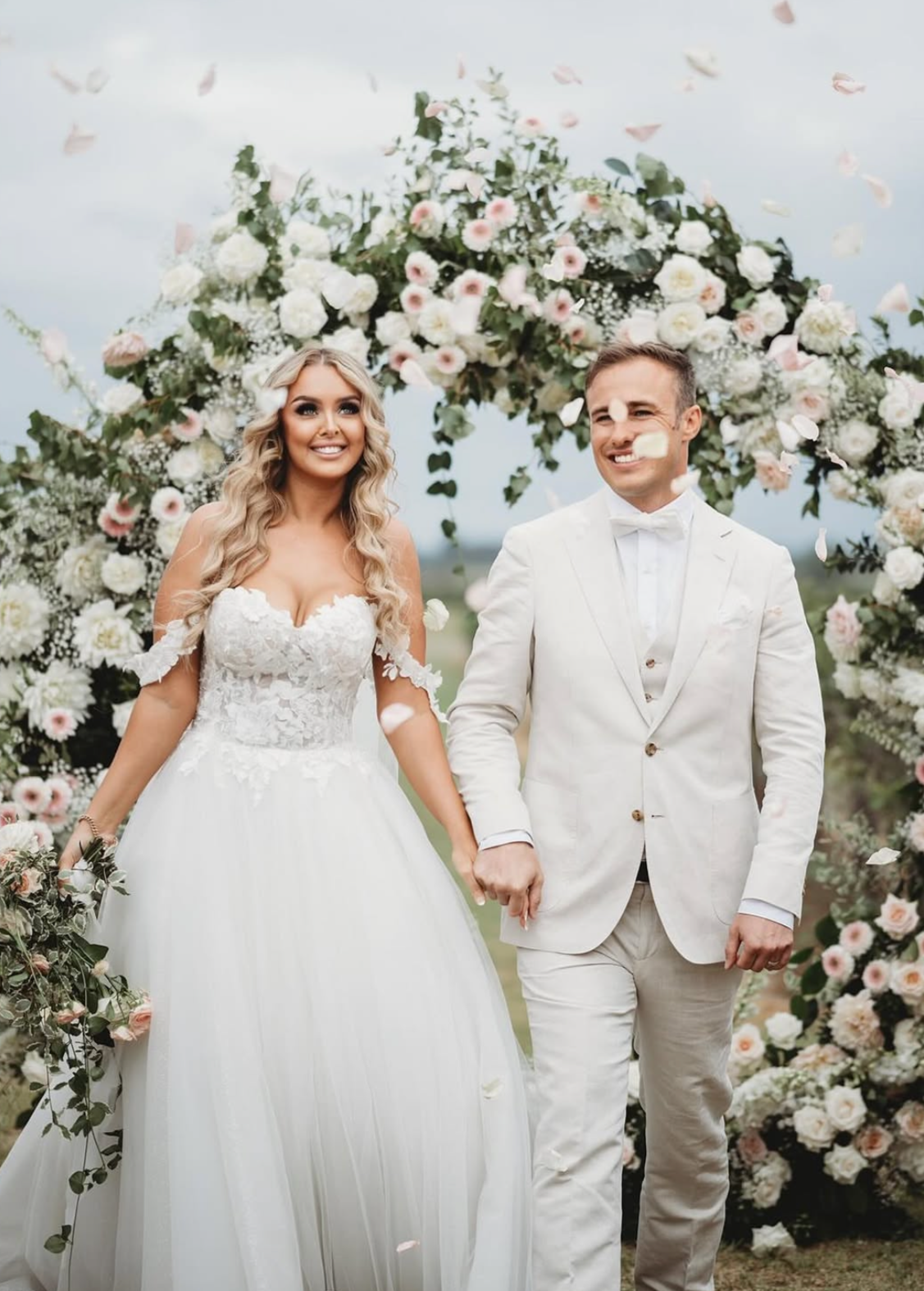 A bride and groom walking hand in hand at their wedding ceremony, surrounded by a floral arch with white and light pink flowers, outdoors on a cloudy day.
