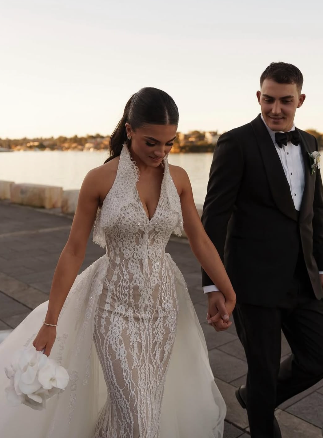 Bride and groom walking hand in hand outdoors at sunset, with water and distant buildings in the background. The bride wears a lace wedding gown and holds a white bouquet, the groom wears a tuxedo with a bow tie.