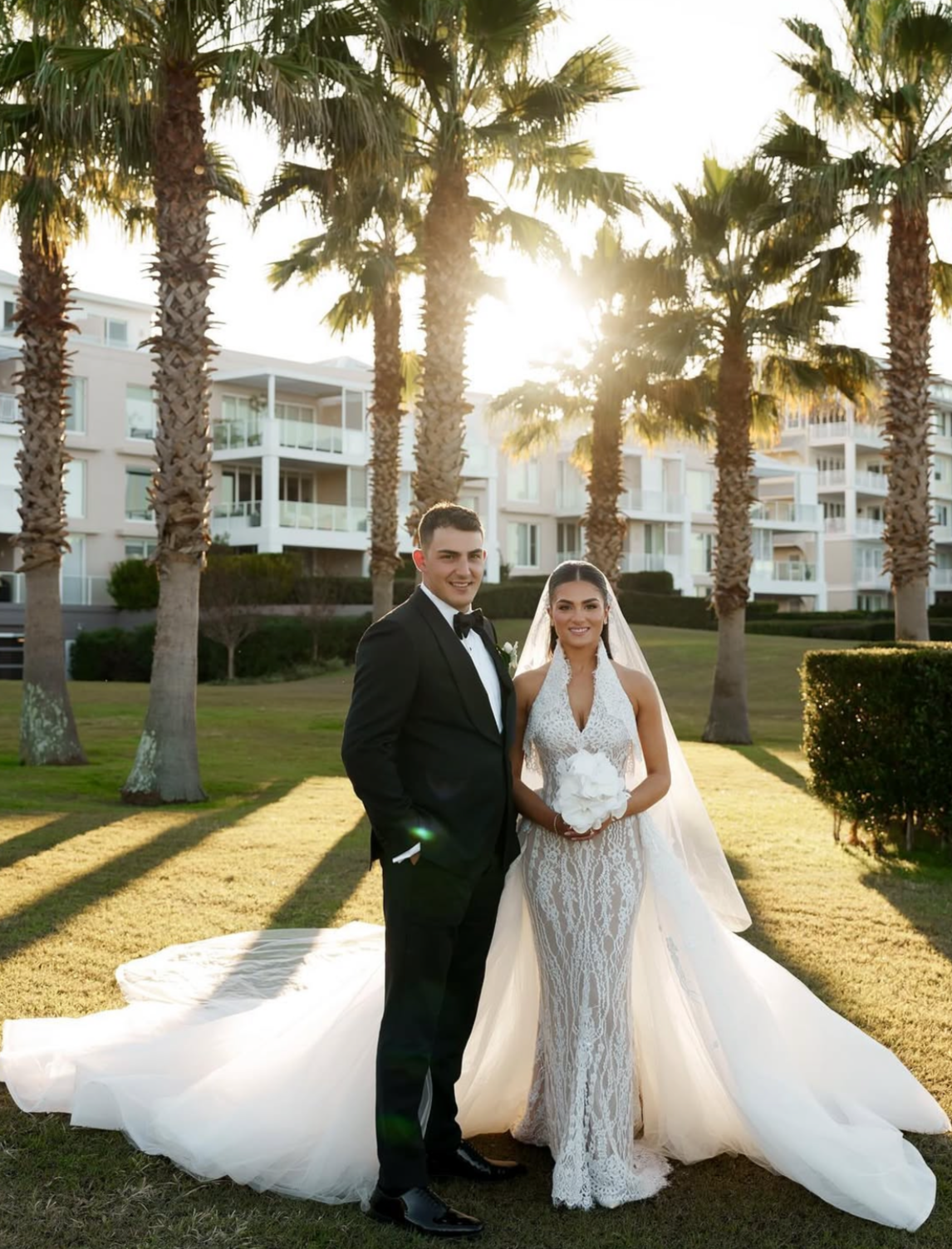 A bride and groom pose for a photo outdoors in front of palm trees and a modern white building at sunset, with lush green grass and a white wedding gown.