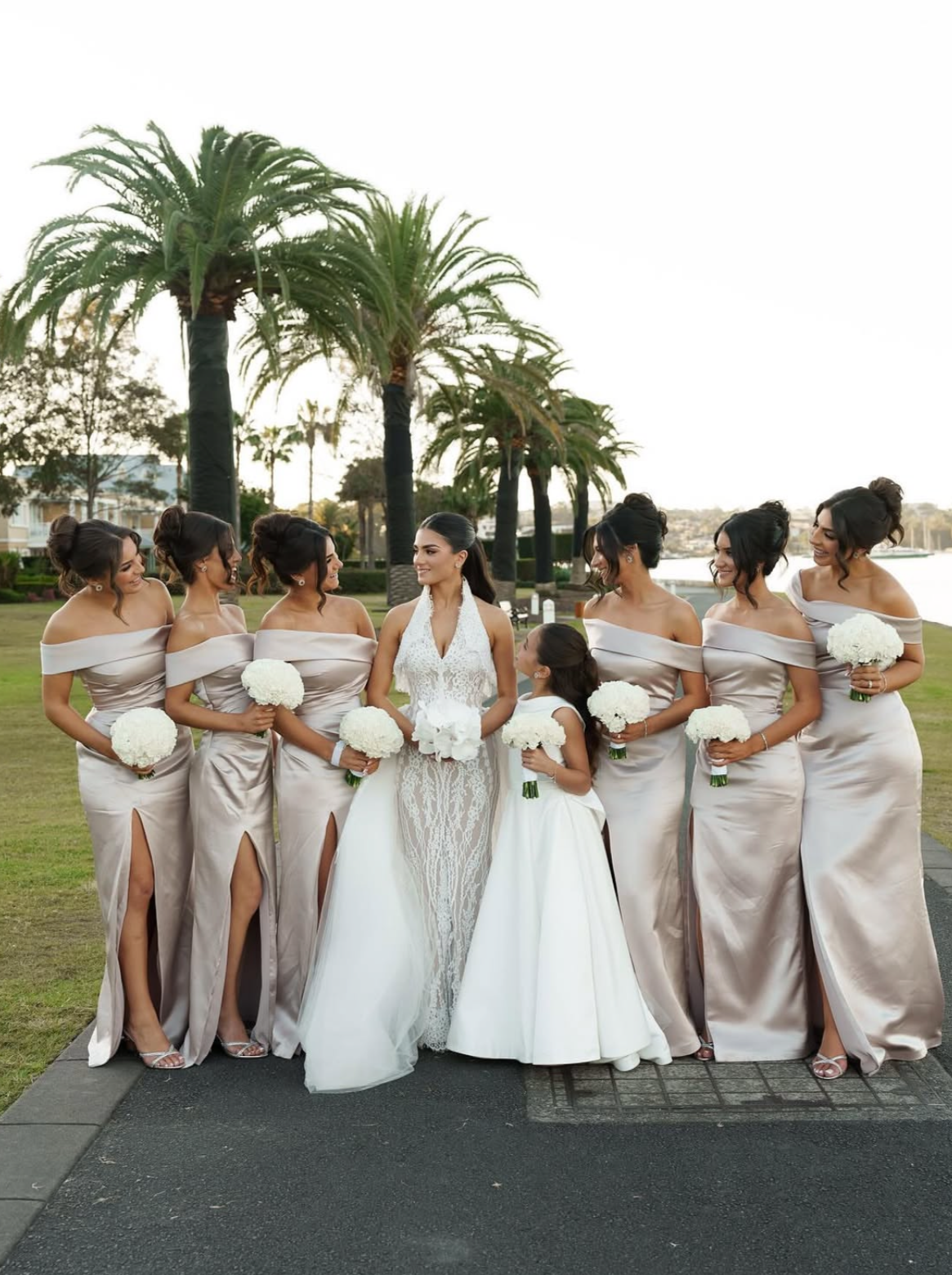 A bride in a white lace wedding gown surrounded by eight bridesmaids in matching off-the-shoulder satin dresses, all holding white bouquets, standing outdoors near palm trees by a waterfront.