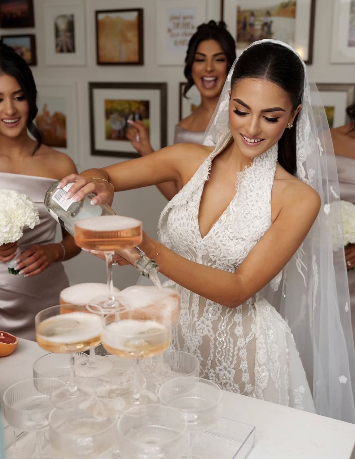 A bride in a white lace wedding dress with a veil pours pink champagne into a tower of coupe glasses, surrounded by bridesmaids holding white bouquets and smiling, with framed pictures on the wall in the background.
