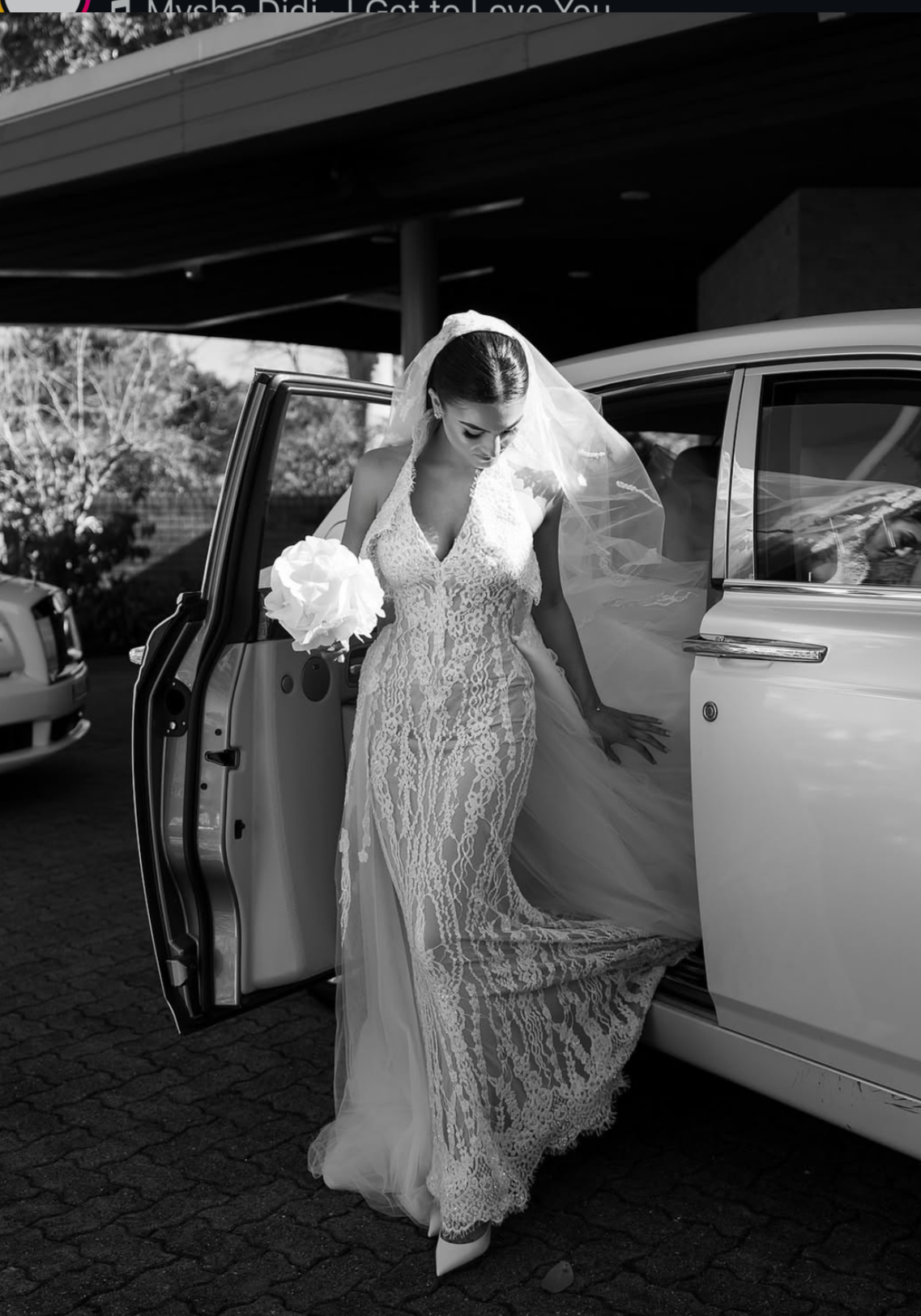 A bride in a lace wedding gown with a veil, holding a bouquet, stepping out of a luxury car.