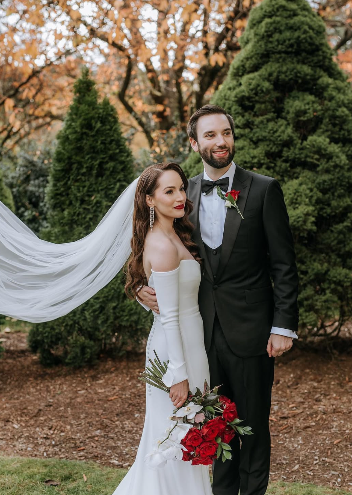A bride and groom stand together outdoors during fall, with trees displaying autumn leaves in the background. The bride is in a white off-shoulder wedding gown with a flowing veil, holding a bouquet of red and white flowers. The groom is in a black t