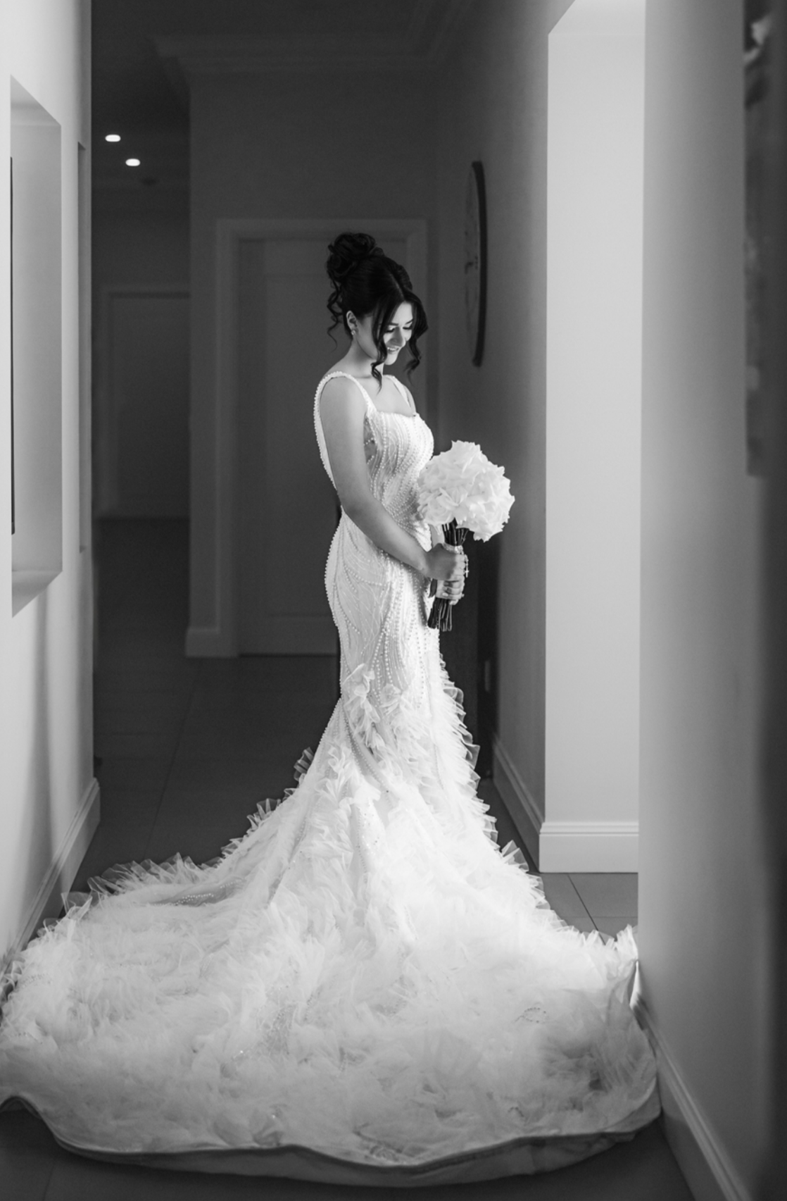 A bride in a detailed wedding gown holding a bouquet of flowers, standing in a hallway with light-colored walls and a clock on the wall.