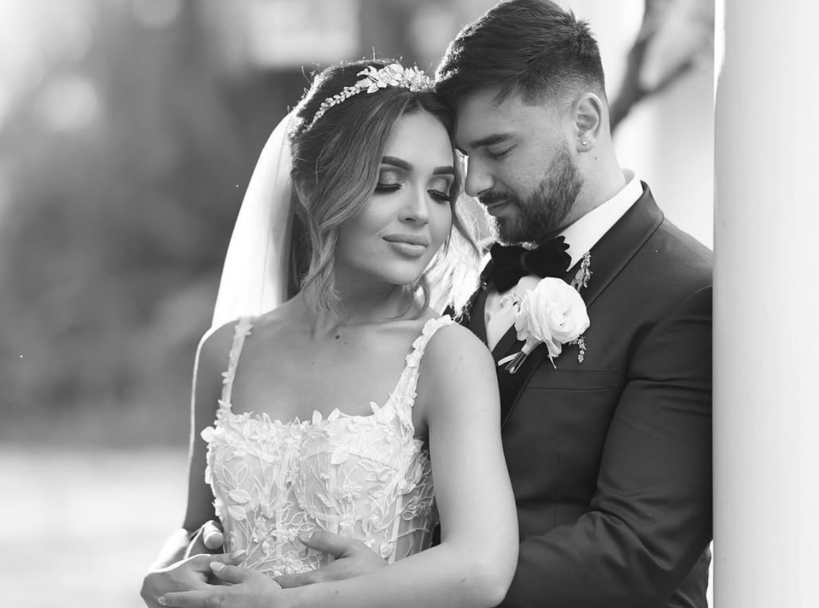 A bride and groom in wedding attire, embracing outdoors, with the groom leaning his forehead against the bride's, both with eyes closed, in a black-and-white photo.