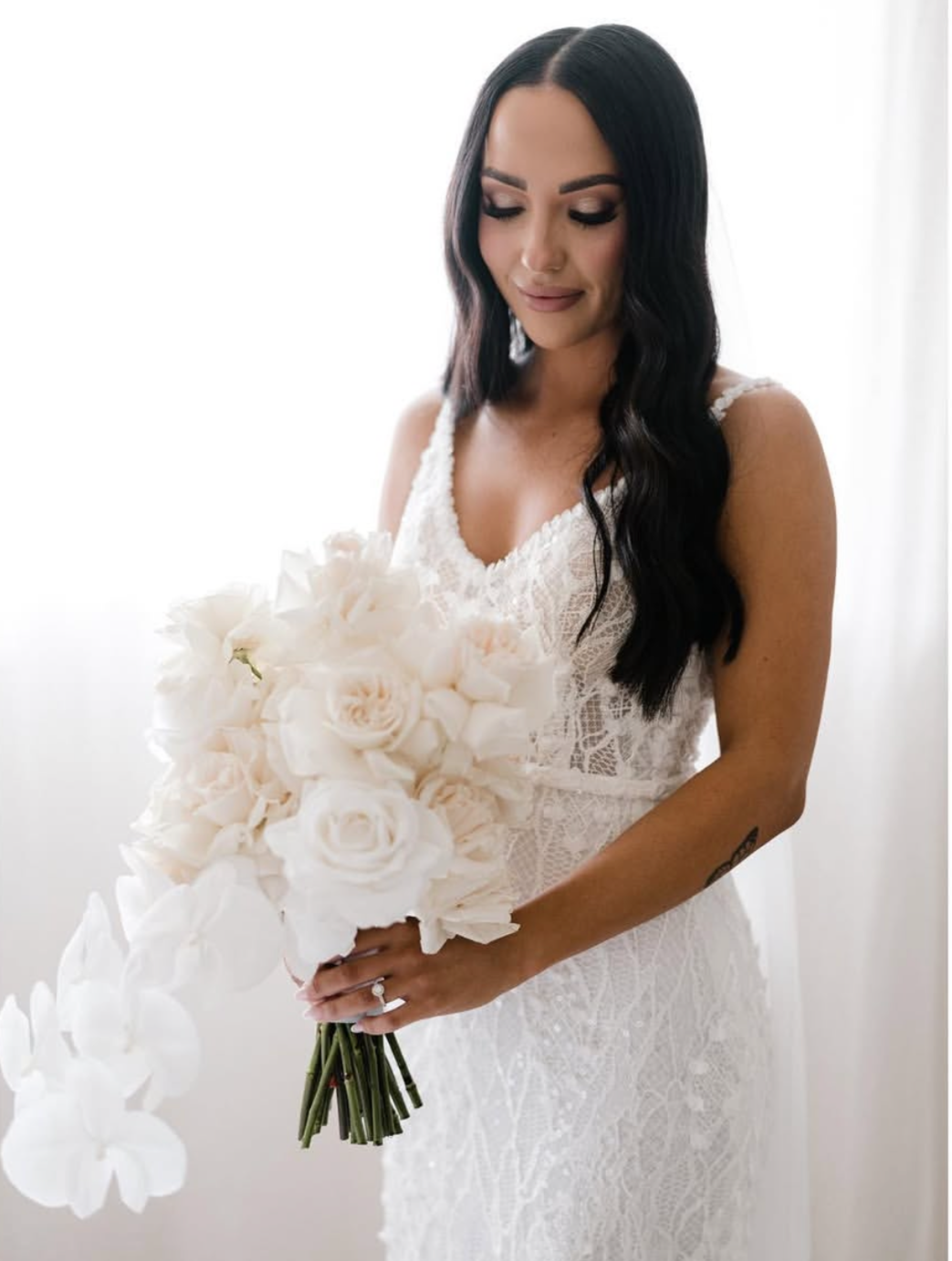 A woman in a white lace dress holding a bouquet of white roses and orchids, with dark wavy hair, standing near a bright window.