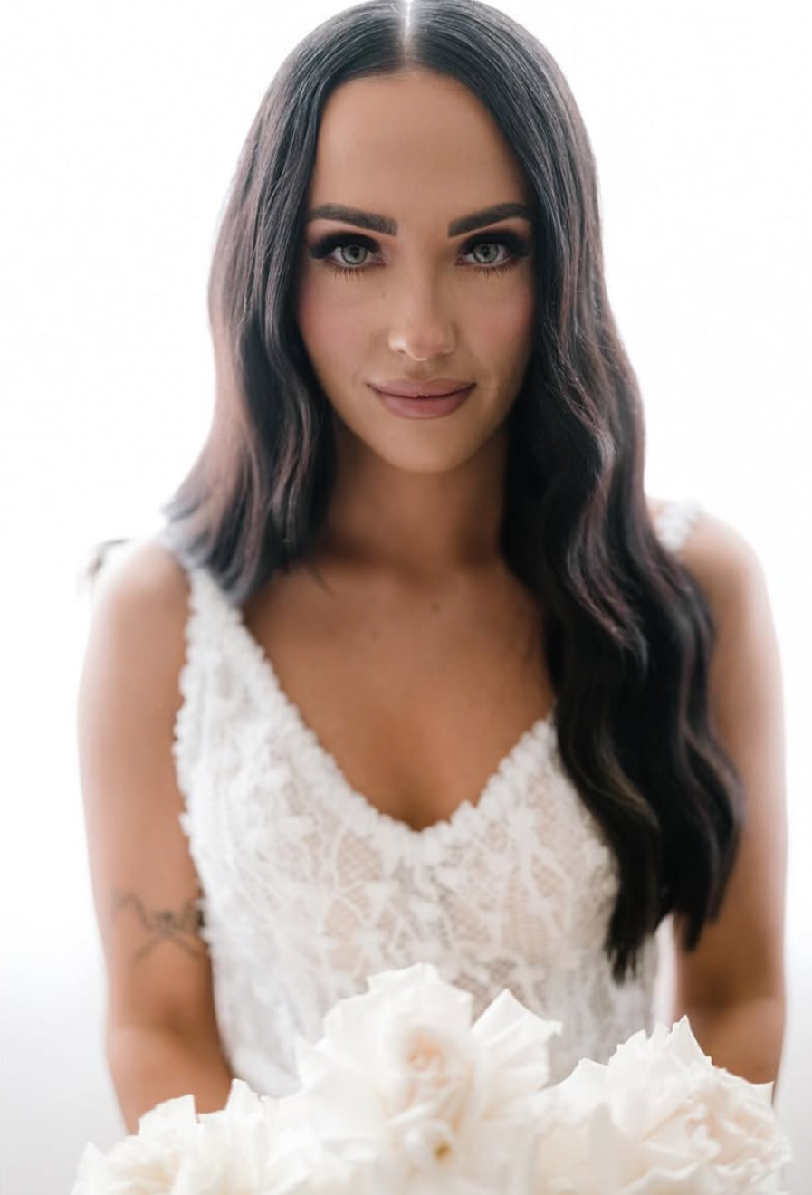 A woman with long dark wavy hair, blue eyes, and makeup, wearing a white lace dress, holding a bouquet of white flowers, smiling softly.