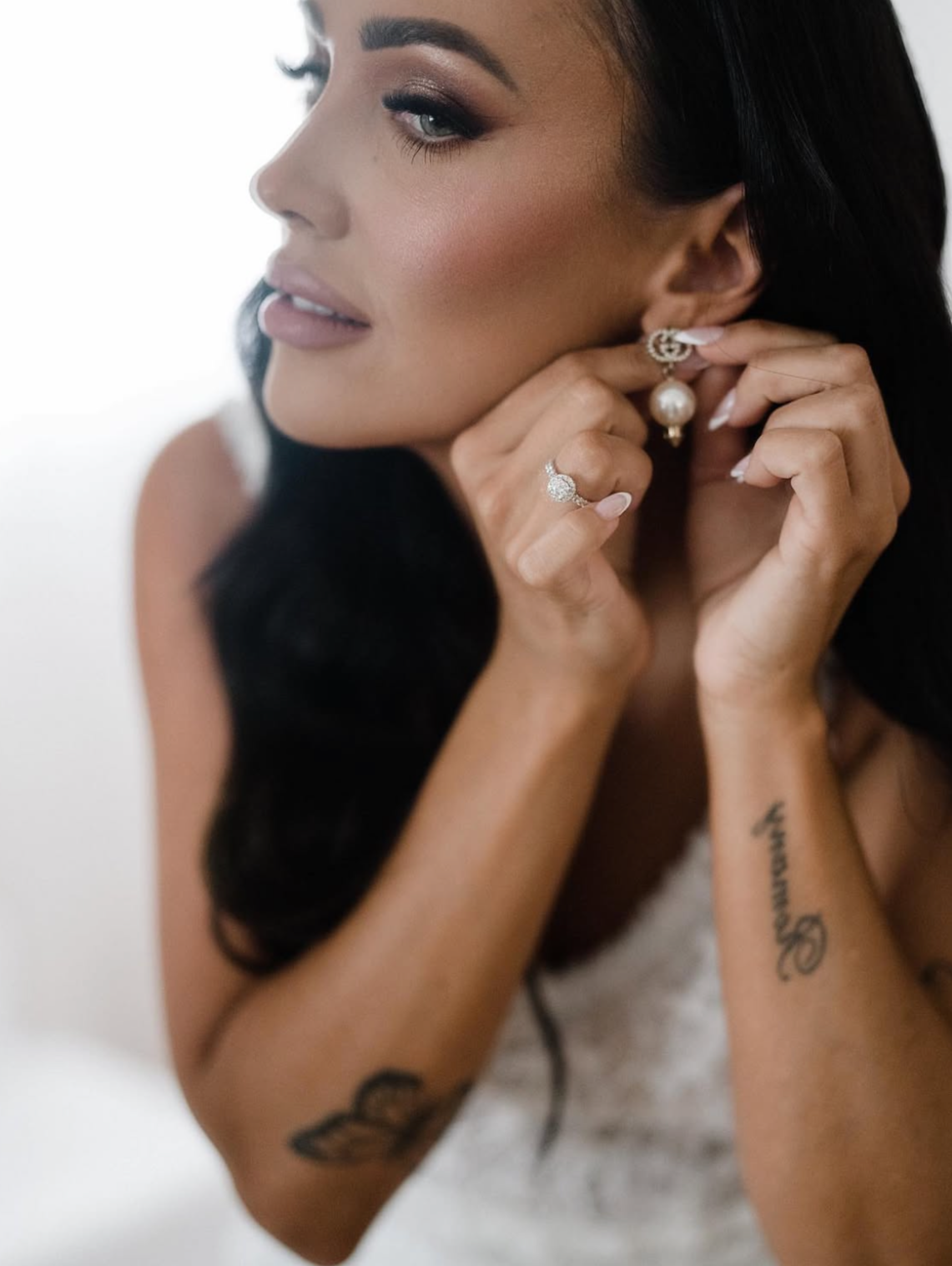 A woman with dark hair and makeup is putting on earrings, wearing a ring on her finger, and has tattoos on her arms.