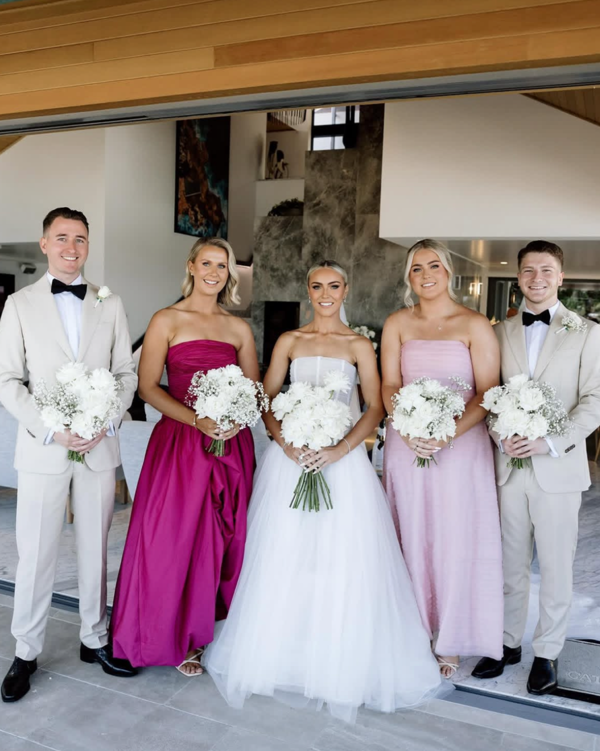 A group of five people, two men and three women, dressed in formal attire at a wedding. The women are wearing pink and white dresses, and the men are in light-colored suits with black bow ties. They are holding bouquets of white flowers and are stand