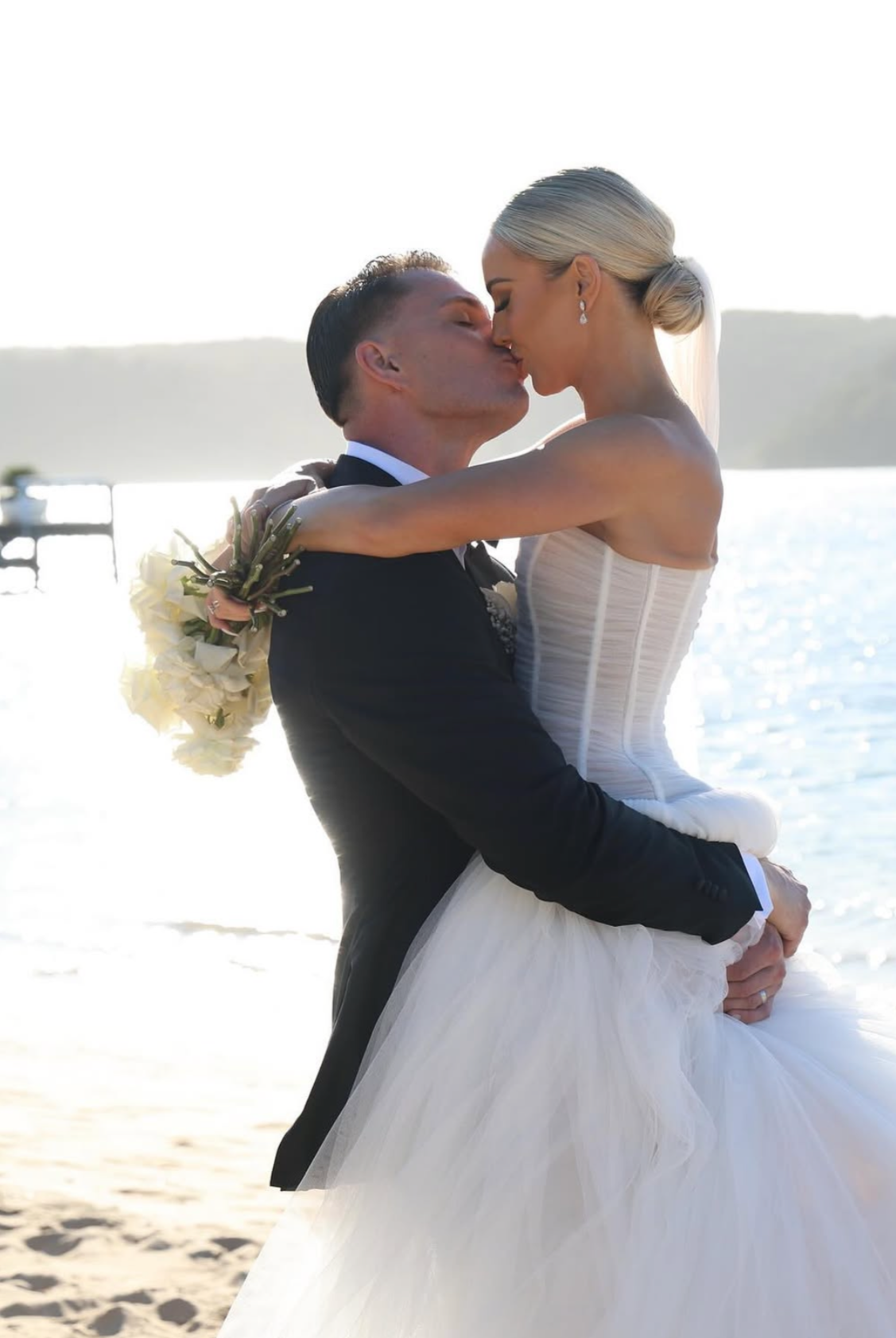 A bride and groom share a kiss by the water, with the bride wearing a white strapless wedding dress and the groom in a tuxedo, during sunset.