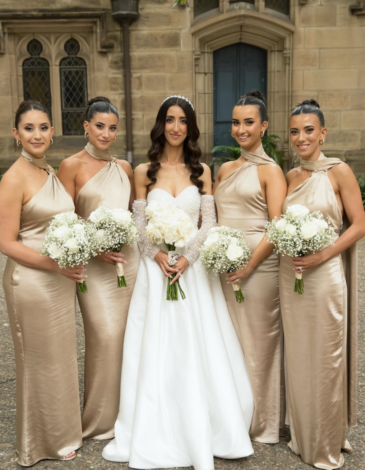 A bride in a white wedding gown with dark hair and long curls, wearing a tiara, is standing in the center of five bridesmaids in matching champagne-colored dresses, all holding white bouquets, in front of an old stone building with Gothic-style windo