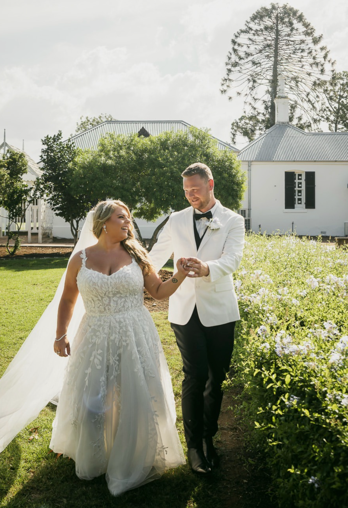 A newlywed couple holding hands and smiling in a garden, with a white house, green trees, and a cloudy sky in the background.
