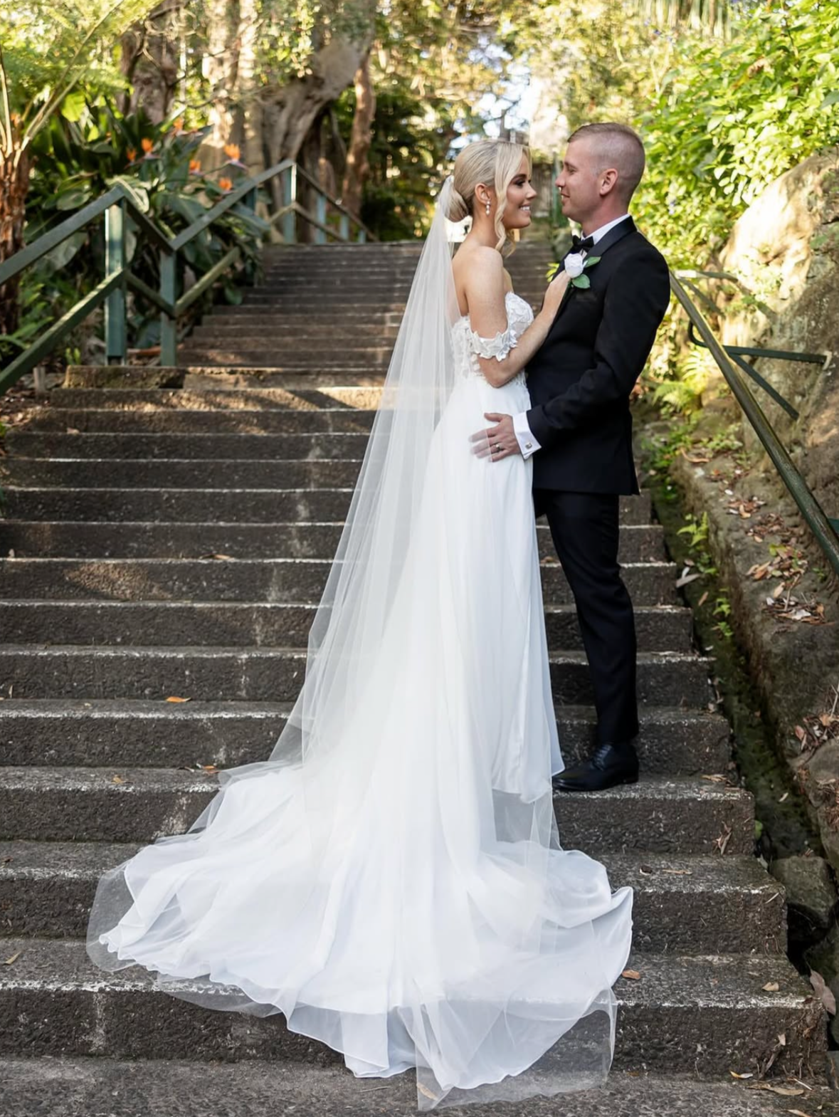 A bride and groom stand on outdoor stone stairs, smiling at each other during their wedding. The bride wears a white wedding gown with a long veil, and the groom is in a black tuxedo. Green foliage and trees surround them.