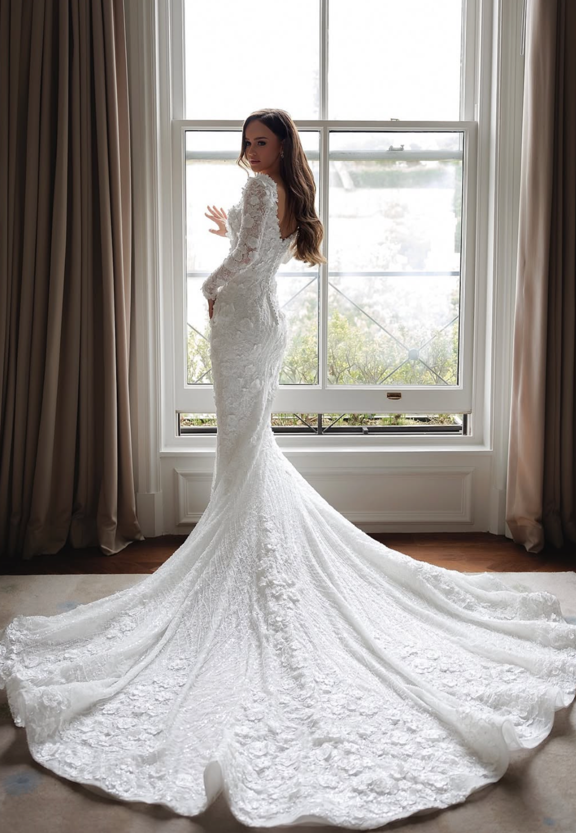 A woman in a white lace bridal gown with a train stands indoors near a large window, looking over her shoulder with her hand touching the window frame.