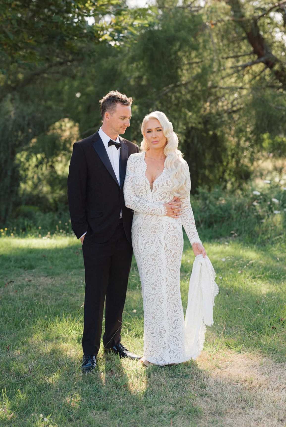 A bride and groom stand outdoors on a grassy area, with the groom in a black tuxedo and the bride in a long white lace dress holding a white cloth, surrounded by green trees and sunlight.