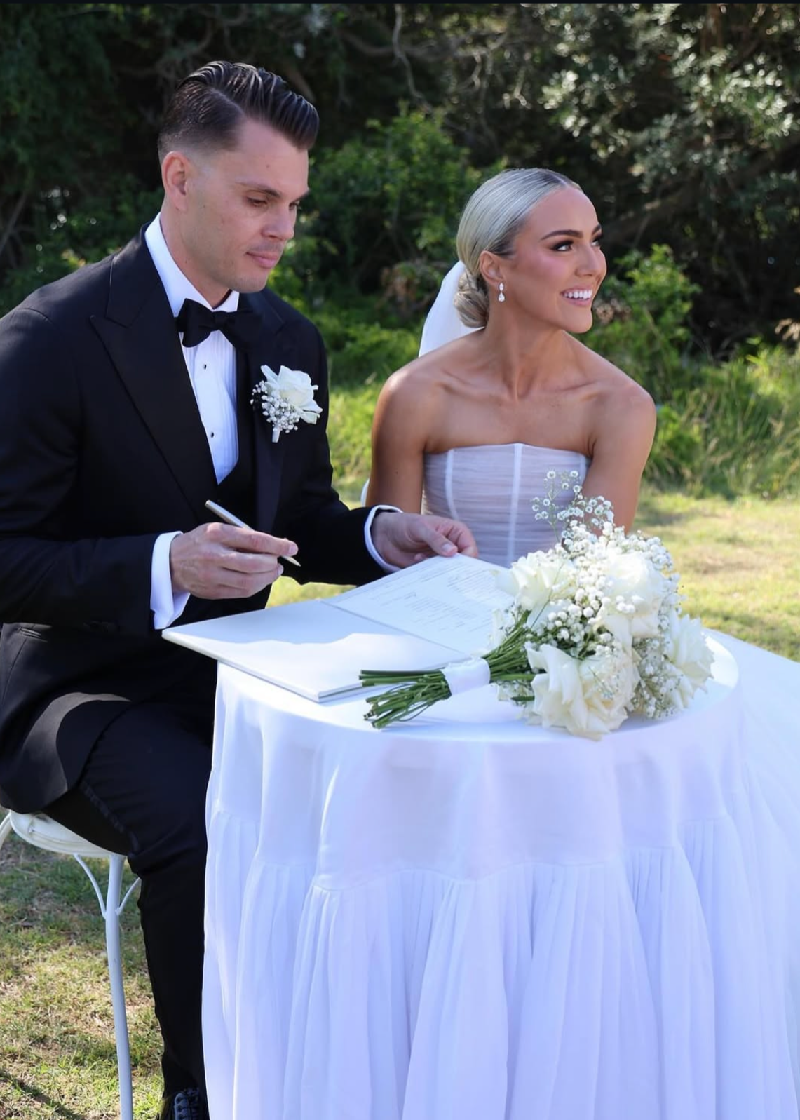 A bride and groom signing marriage documents outdoors during a wedding ceremony, with a bouquet of white flowers on the table.