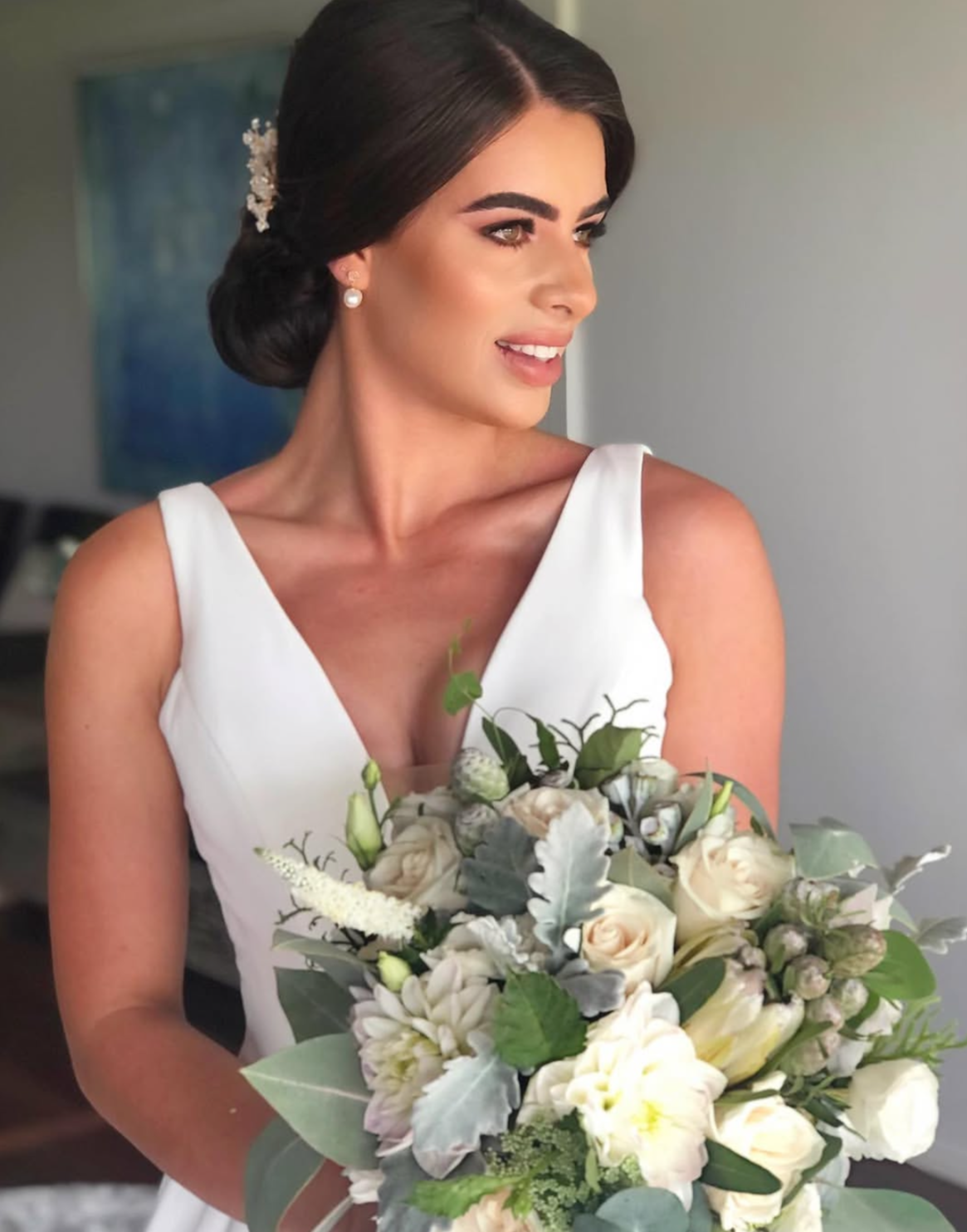 A woman in a white dress holding a large bouquet of white and green flowers, with her dark hair styled in an elegant updo and decorated with a pearl and floral hairpiece.