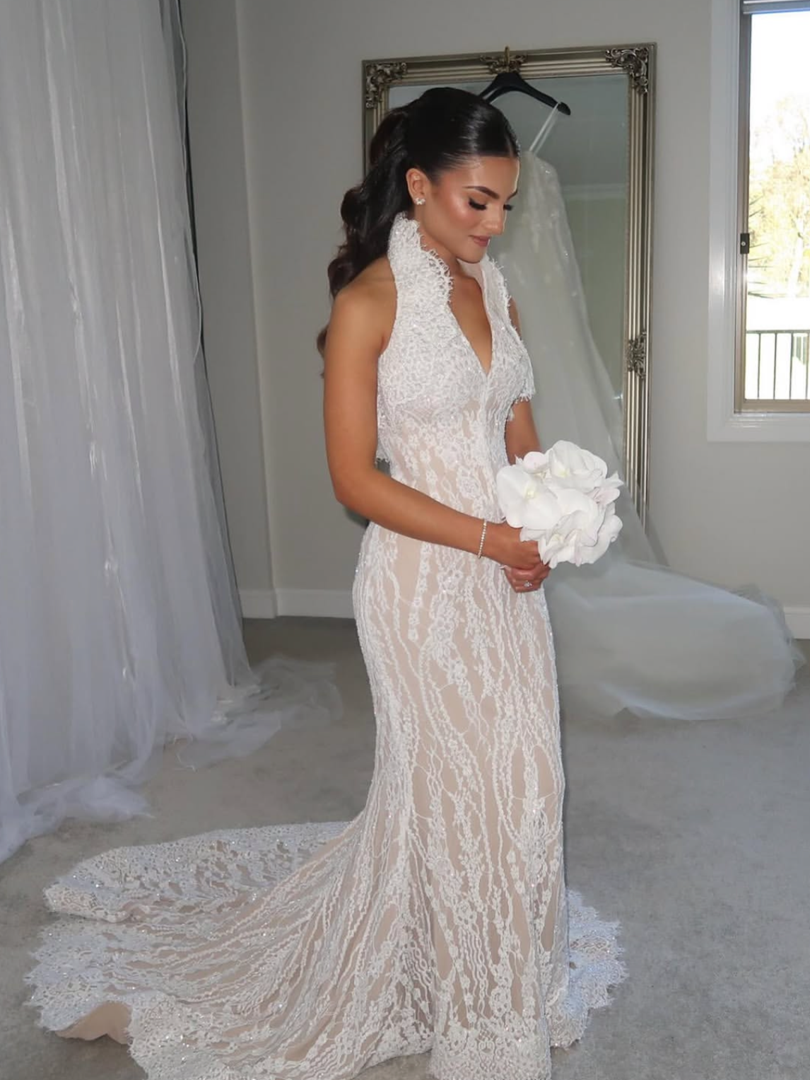 A bride with dark hair styled in loose curls, wearing a fitted white lace wedding gown with a train, holding a bouquet of white flowers, standing indoors near a mirror and a window.