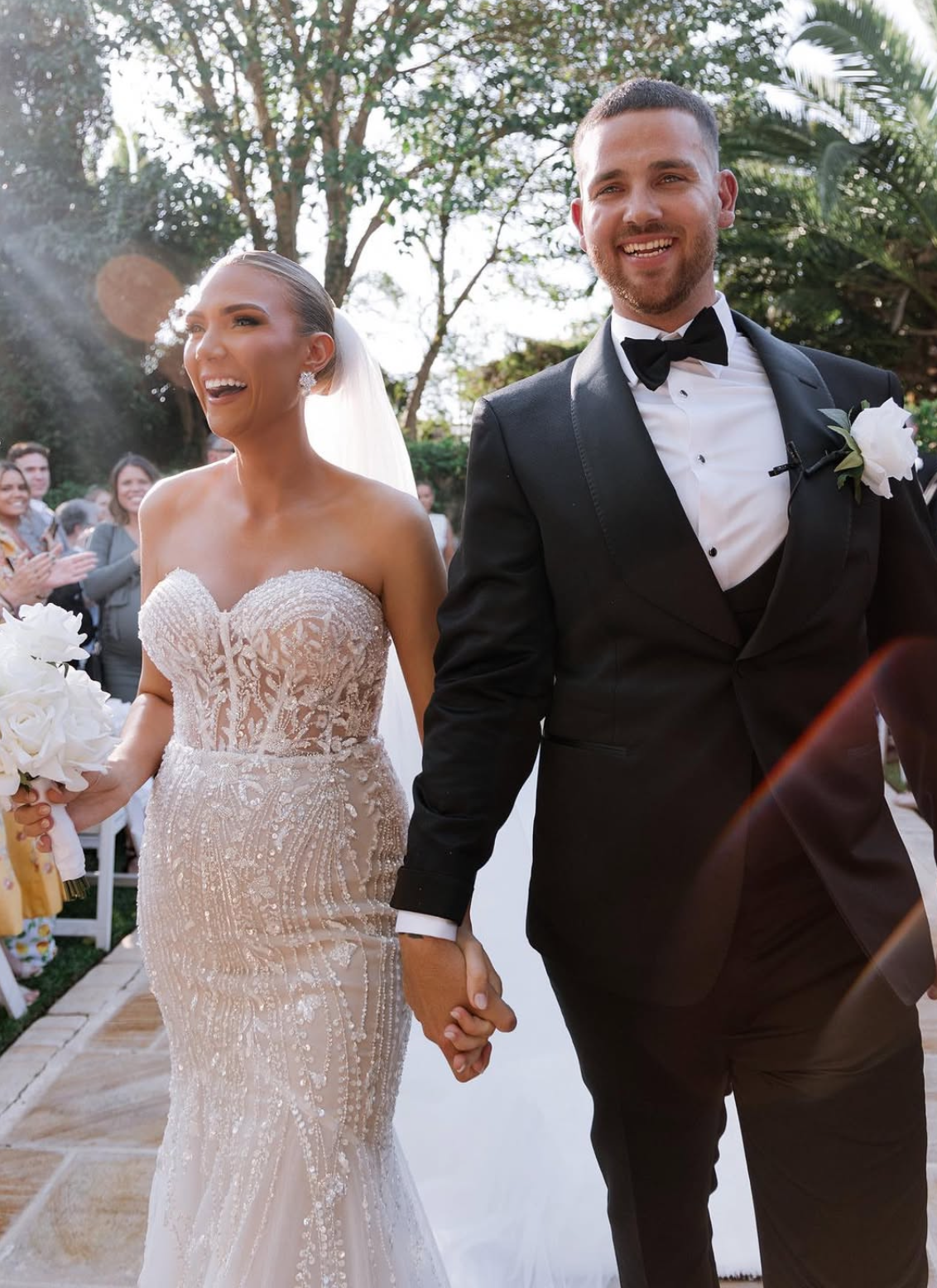 A bride and groom holding hands, smiling, walking outdoors during their wedding ceremony, with guests and trees in the background.