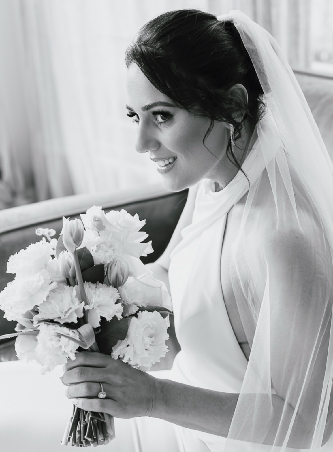 Black and white photo of a smiling bride holding a bouquet of flowers, sitting on a bench, wearing a wedding dress with a veil.
