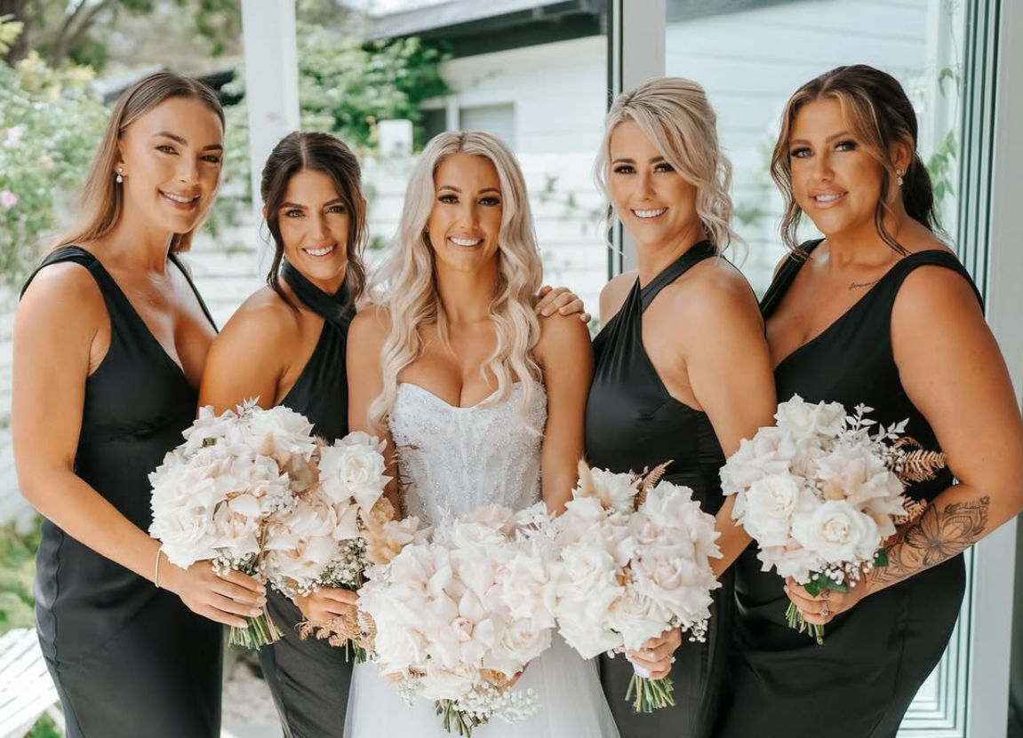Bride surrounded by five bridesmaids, all smiling and holding bouquets in a well-lit room with large windows.
