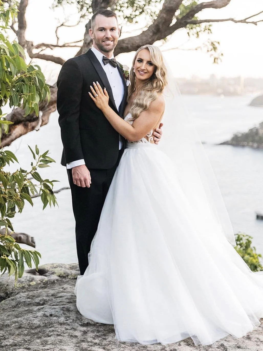 A newlywed couple stands on a rocky surface by the water, smiling at the camera. The groom wears a tuxedo with a bow tie, and the bride wears a white wedding gown with a long veil. There is a tree with green leaves behind them, and the water and dist