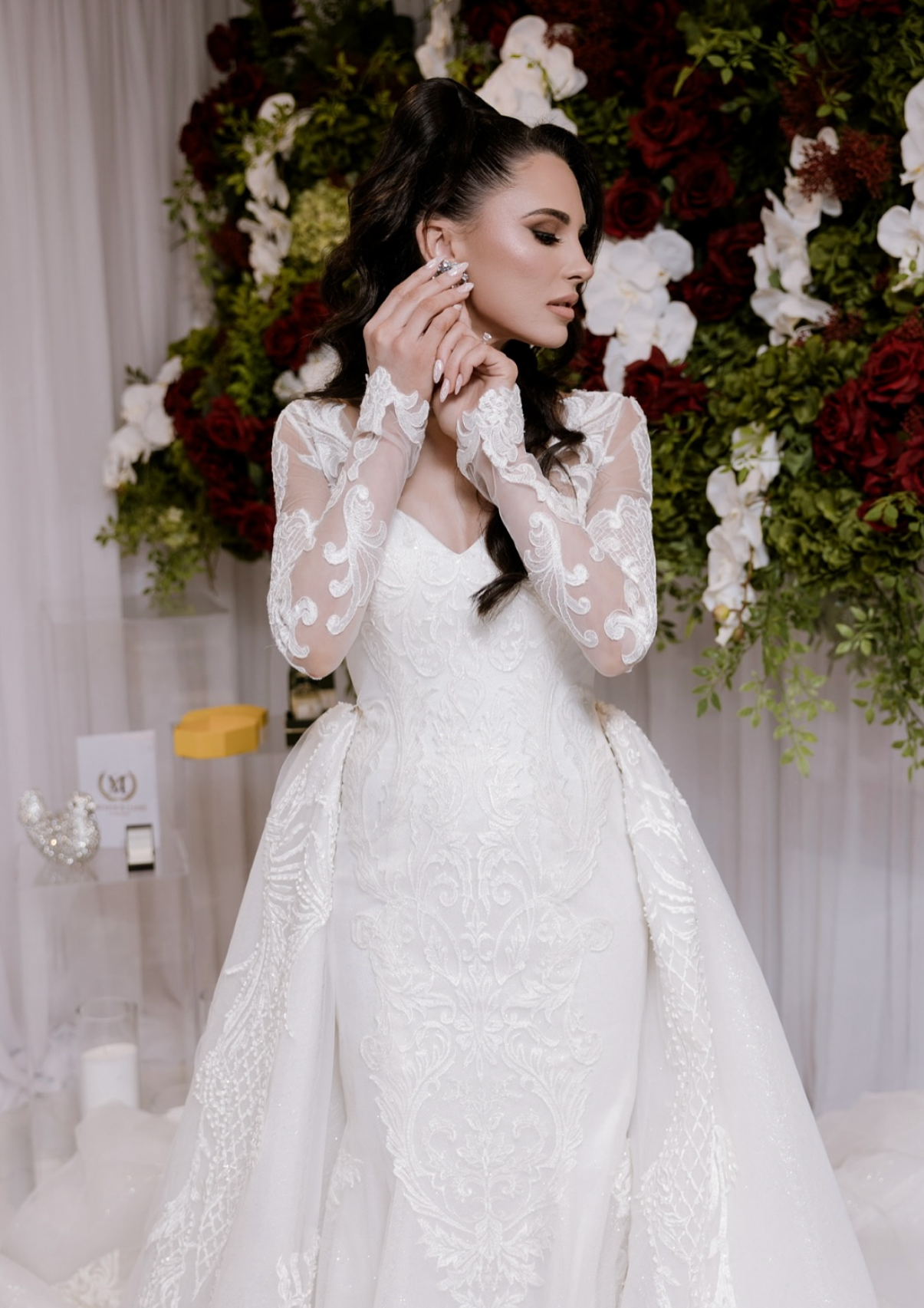 A woman in a wedding dress with lace details and long sleeves, standing in front of a floral backdrop of red and white flowers, as she adjusts an earring.