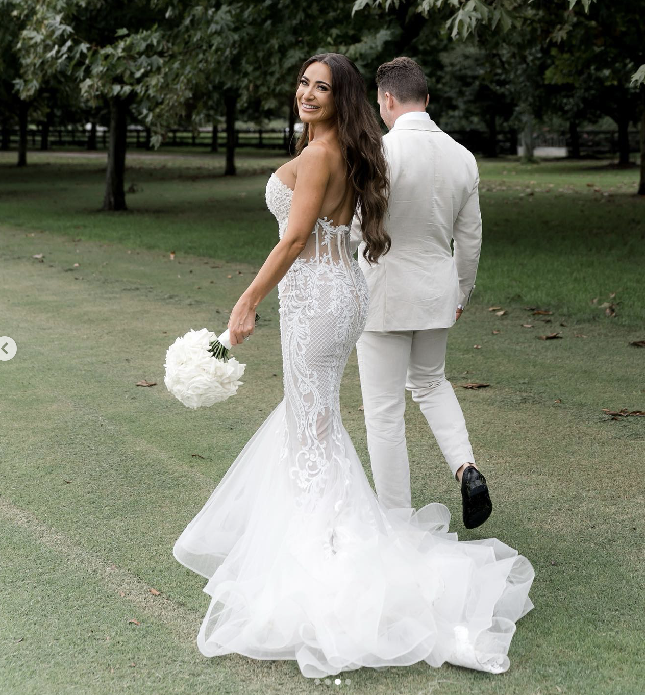 Bride in a lace white wedding gown holding a bouquet of white flowers, smiling and looking over her shoulder, outdoors with a man in a white suit in the background.