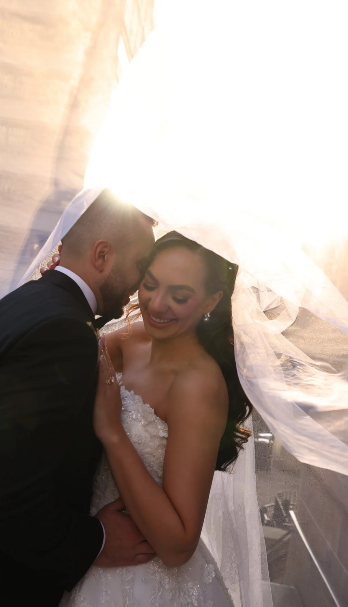 A bride and groom share an intimate moment under a sheer veil, with sunlight illuminating the scene as the groom kisses the bride's forehead.